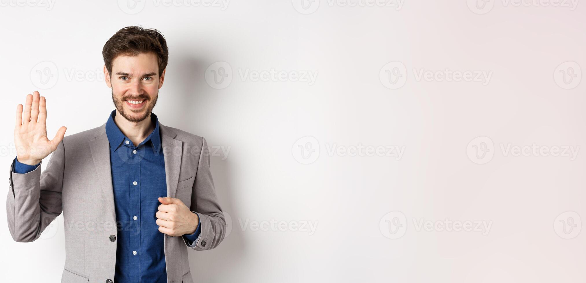 Smiling handsome businessman in suit waving hand, greeting friend with friendly face, say hellp, standing on white background photo
