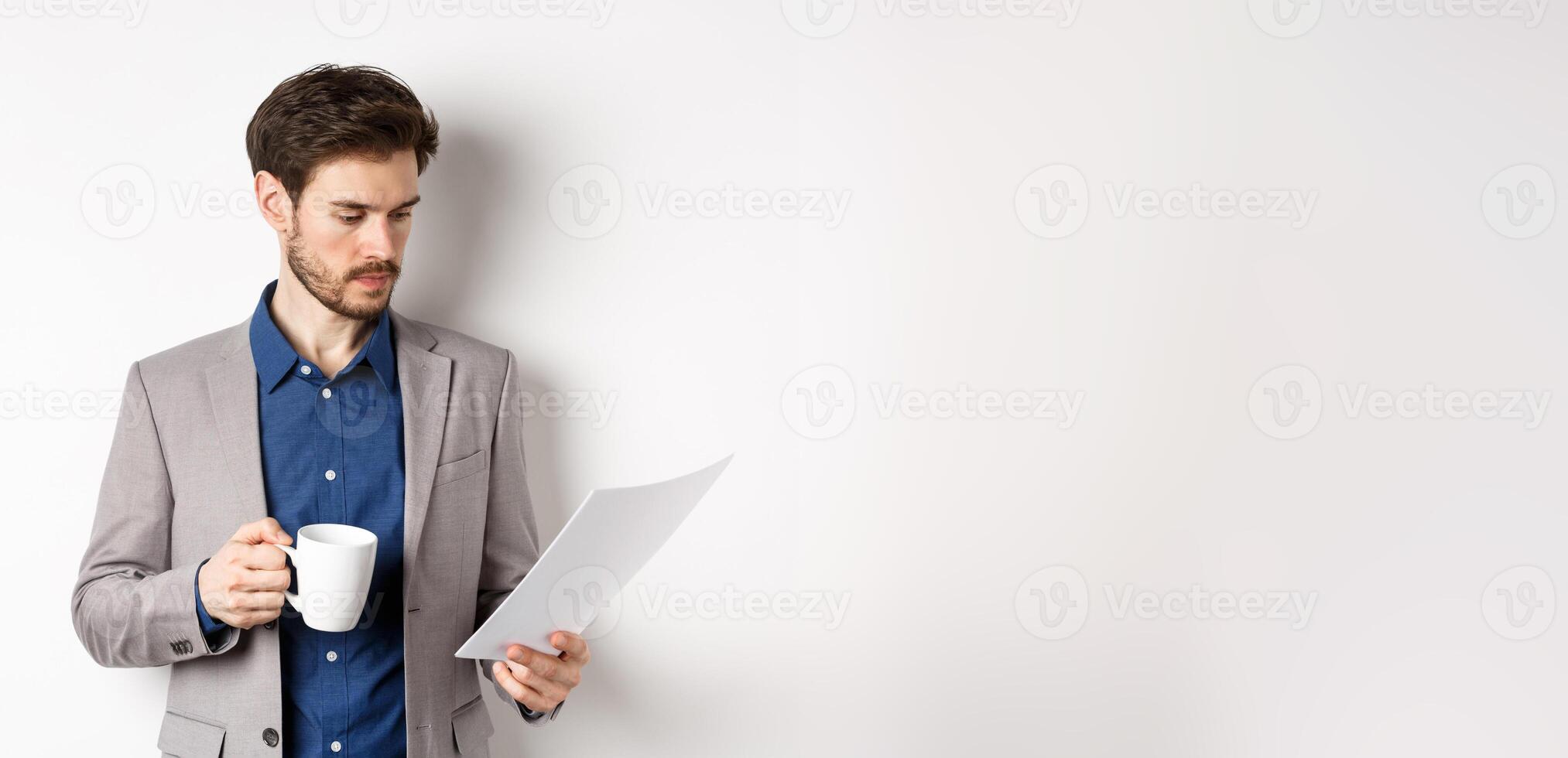 Boss reading documents and drinking morning coffee from mug, looking at paper serious, standing in suit on white background photo
