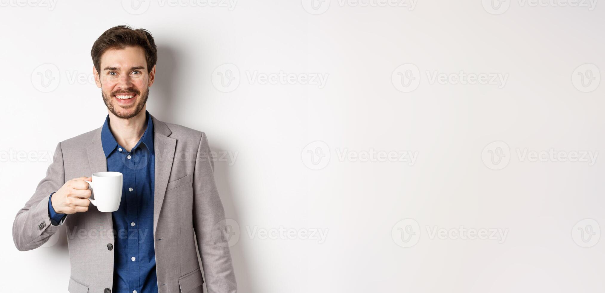 Handsome businessman in suit drinking coffee or tea from office mug, smiling enthusiastic at camera, standing against white background photo
