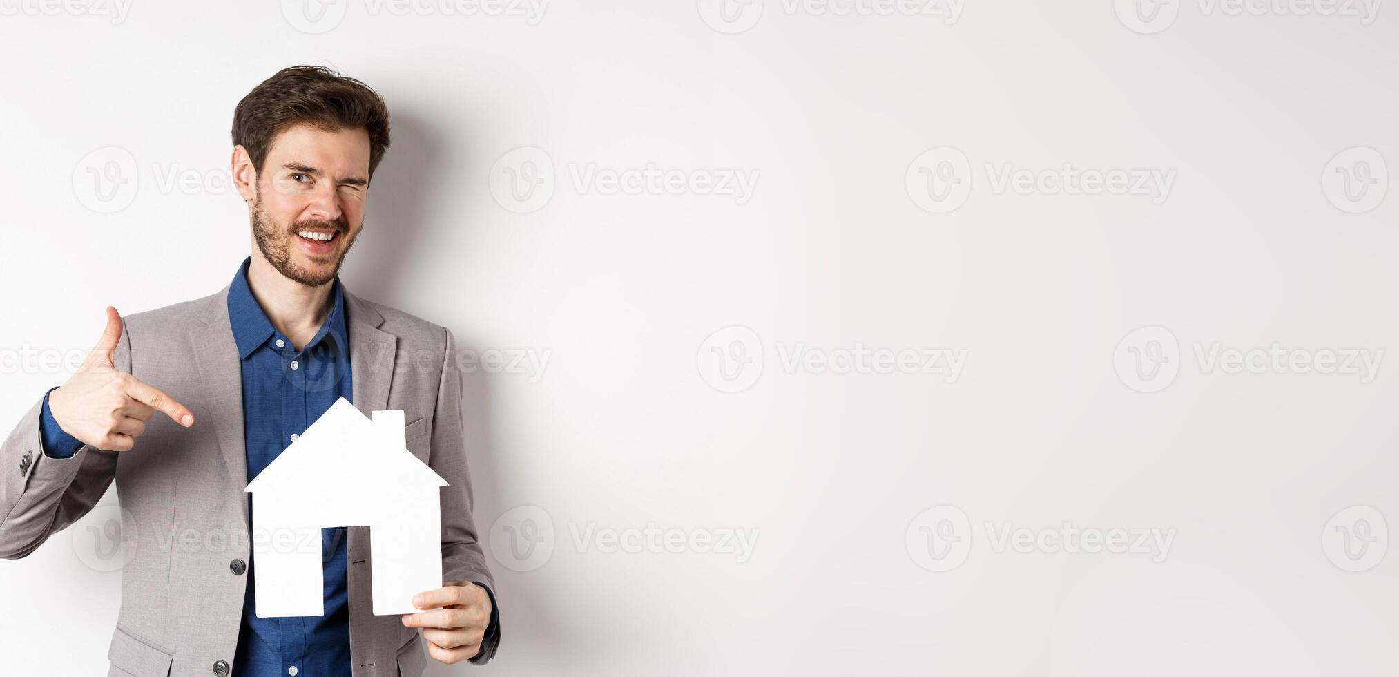 Real estate. Handsome businessman winking and pointing at paper house cutout, advertising company, standing on white background photo