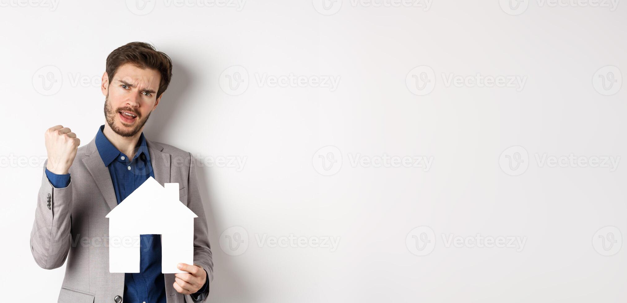 Real estate. Excited man buying house and showing paper home cutout, say yes and fist pump with joy, standing in suit on white background photo