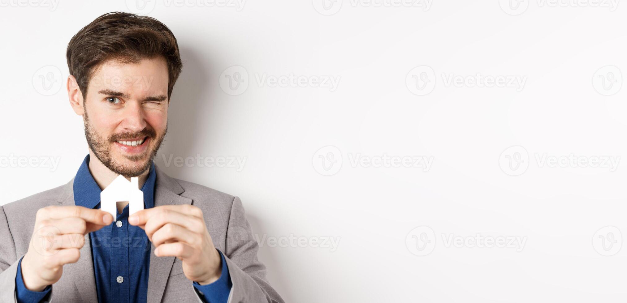 Real estate and insurance concept. Handsome man in suit winking and smiling, showing small paper house cutout, standing on white background photo