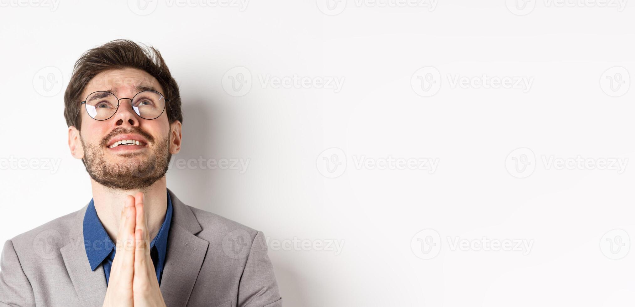 Nervous hopeful man in glasses and suit begging god, asking please and shaking hands in pray, white background photo