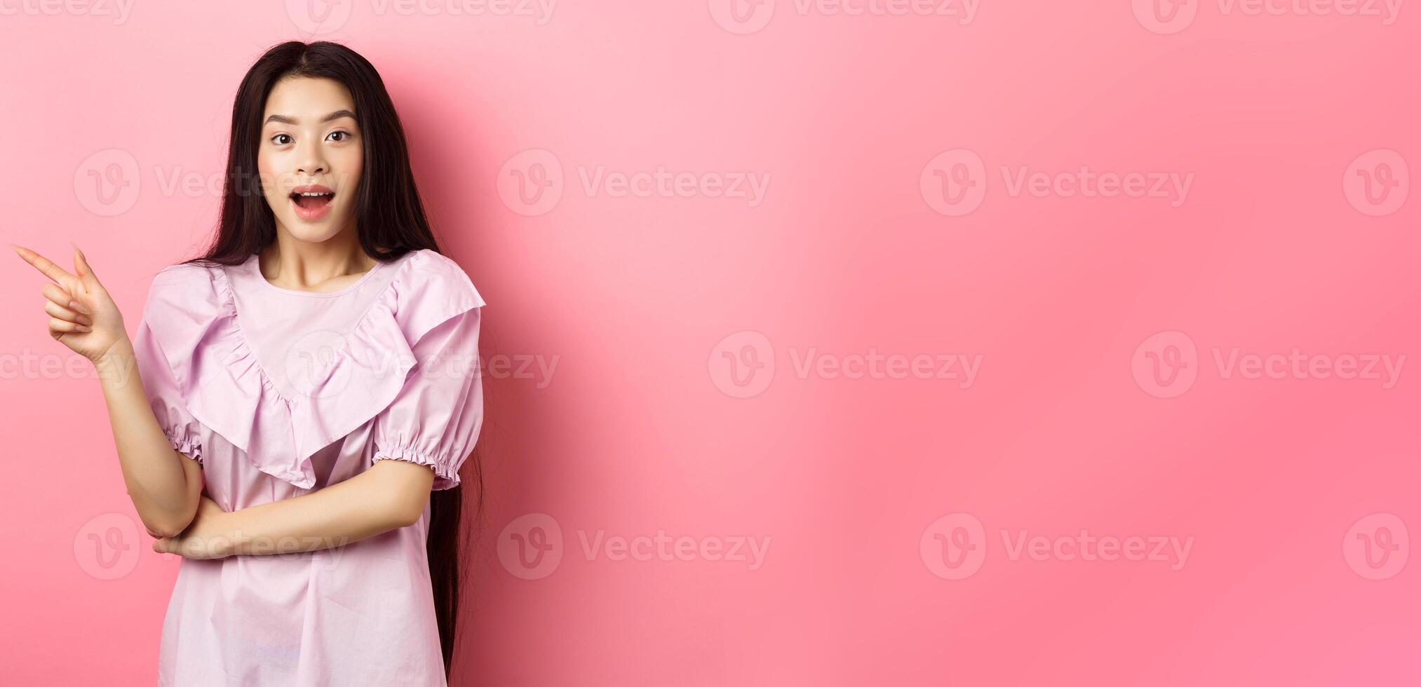 Excited beautiful asian teen girl pointing finger left, open mouth amazed and looking at camera, showing cool promo deal, pink background photo