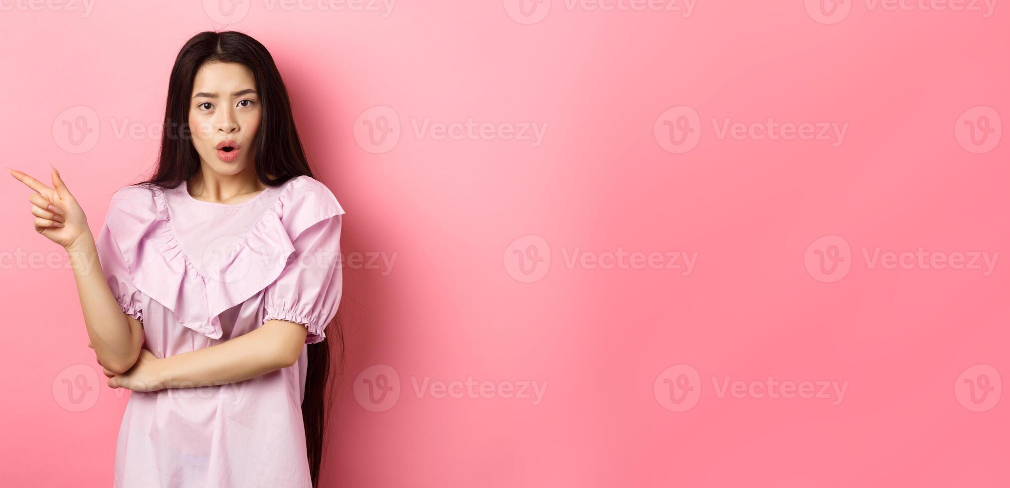 Shocked asian teen girl in dress pointing finger left, gasping wondered and look curious at camera, asking question about promo, standing on pink background photo