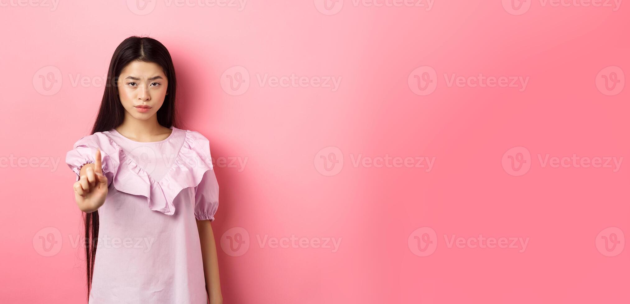 She says no. Serious asian girl shaking finger in stop gesture, prohibit and disagree with person, scolding bad behaviour, standing against pink background photo