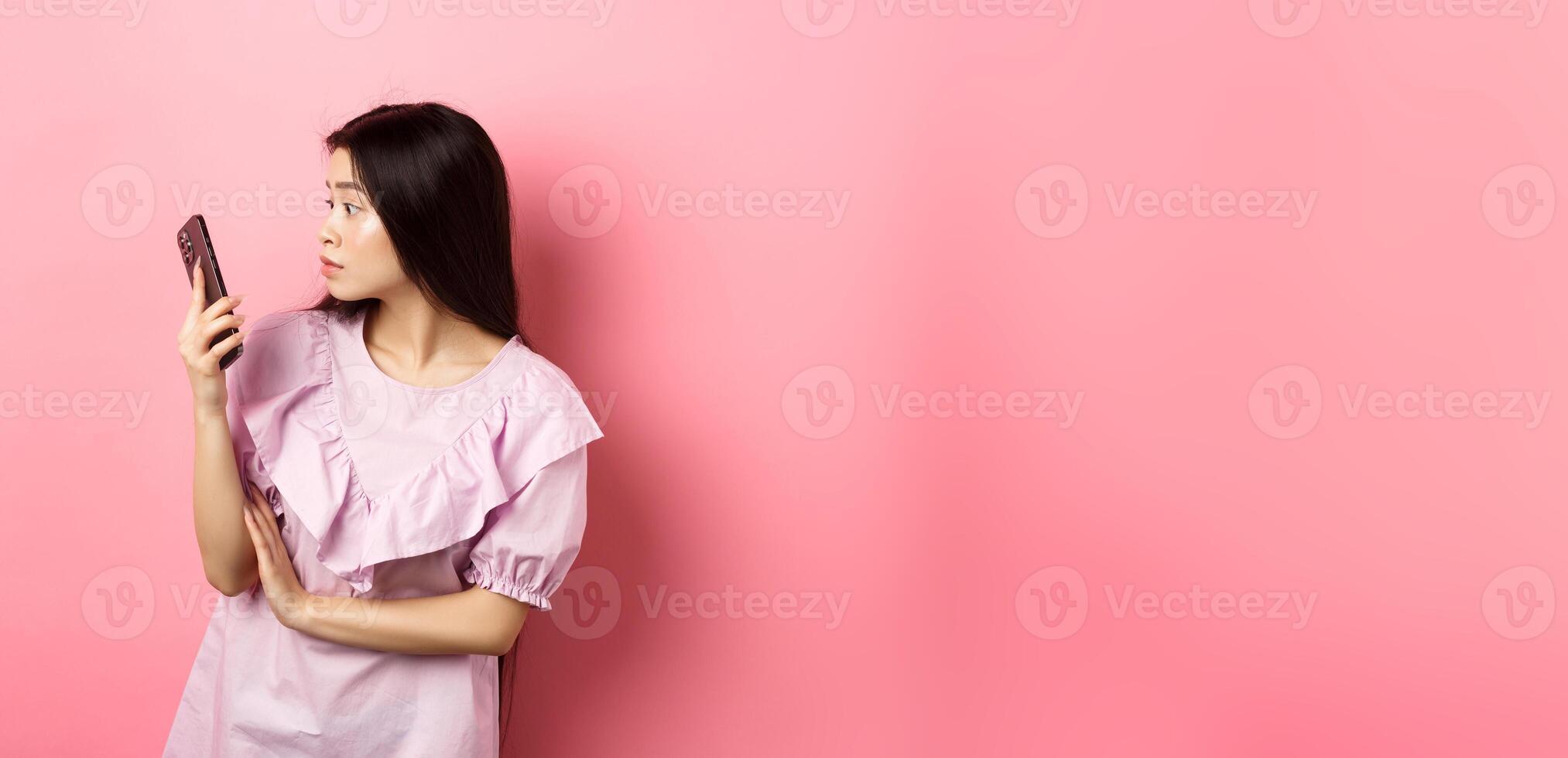 Asian girl look surprised at smartphone screen, reading message, standing in dress against pink background photo