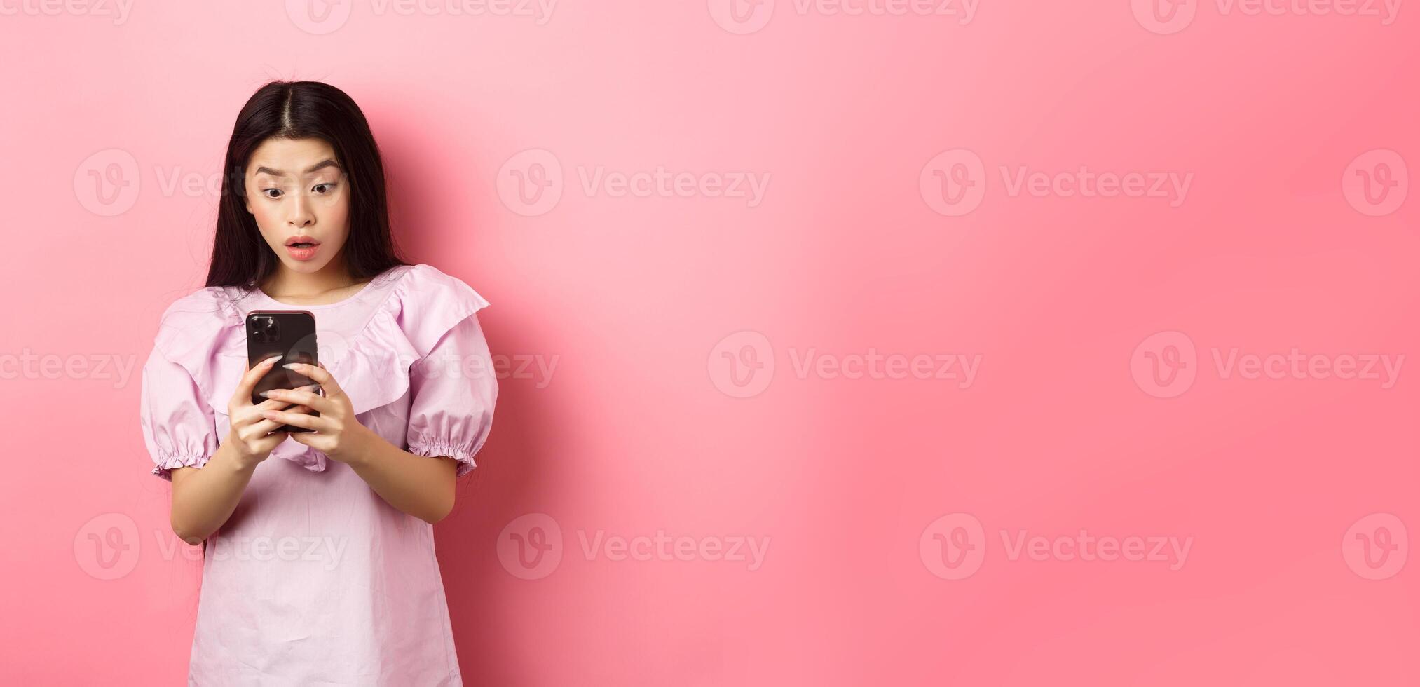 Online shopping. Asian teen girl look surprised at mobile phone screen, reading message with excited expression, standing against pink background photo
