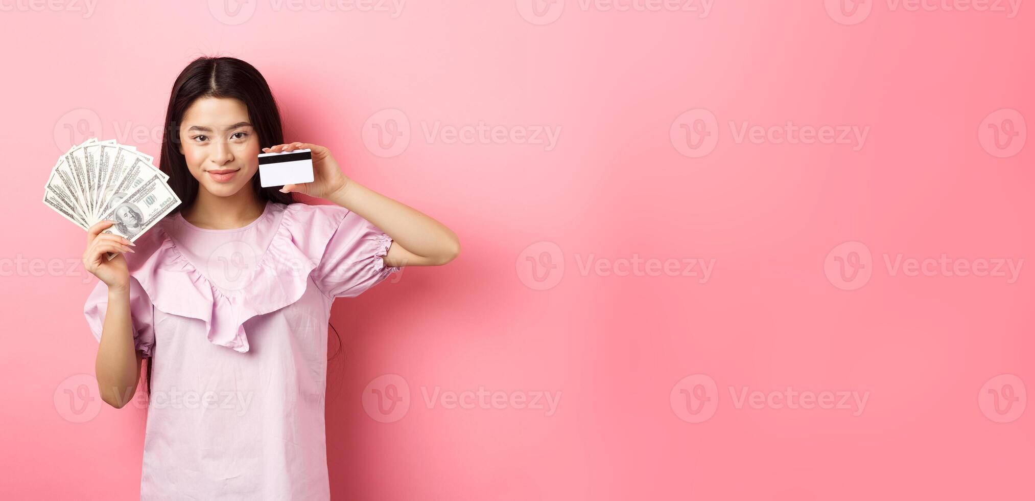 Smiling beautiful asian girl showing money and plastic credit card, giving choice, standing against pink background photo