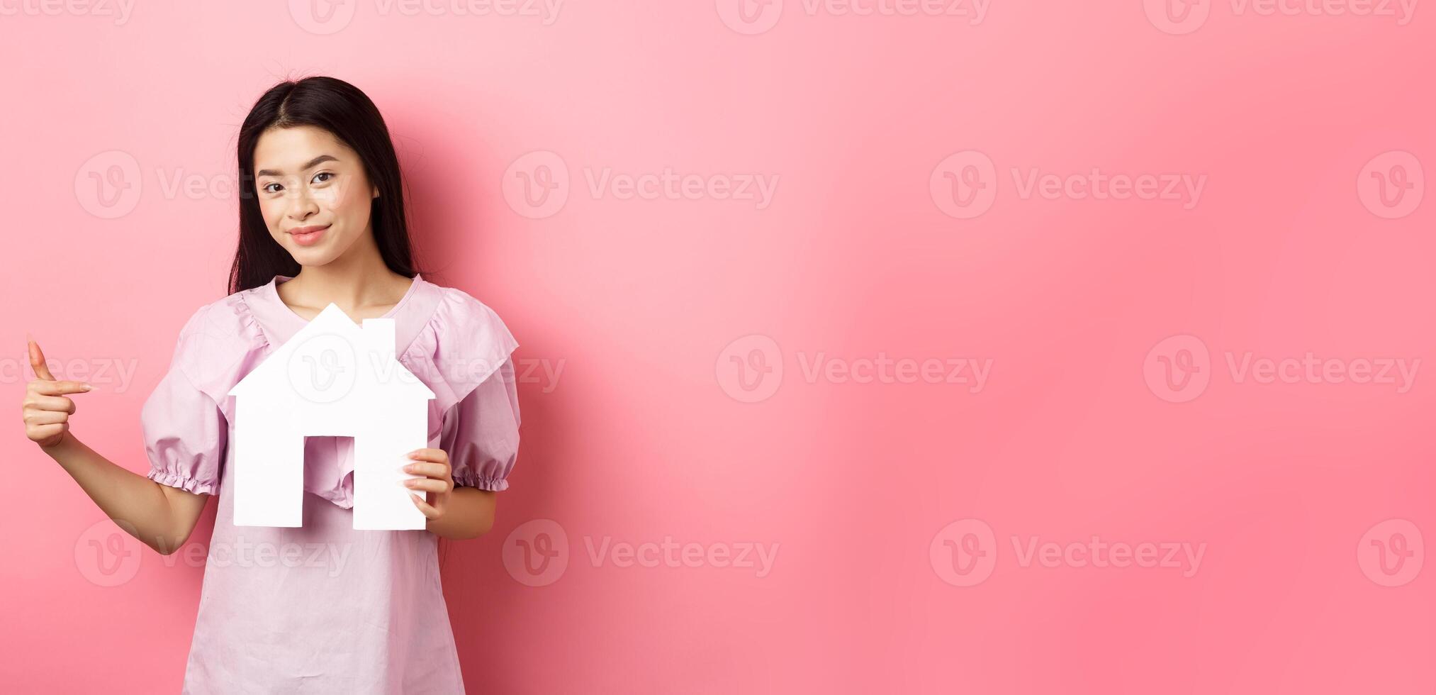 Real estate and insurance concept. Beautiful asian woman pointing at paper house cutout, showing agency logo, standing in dress on pink background photo