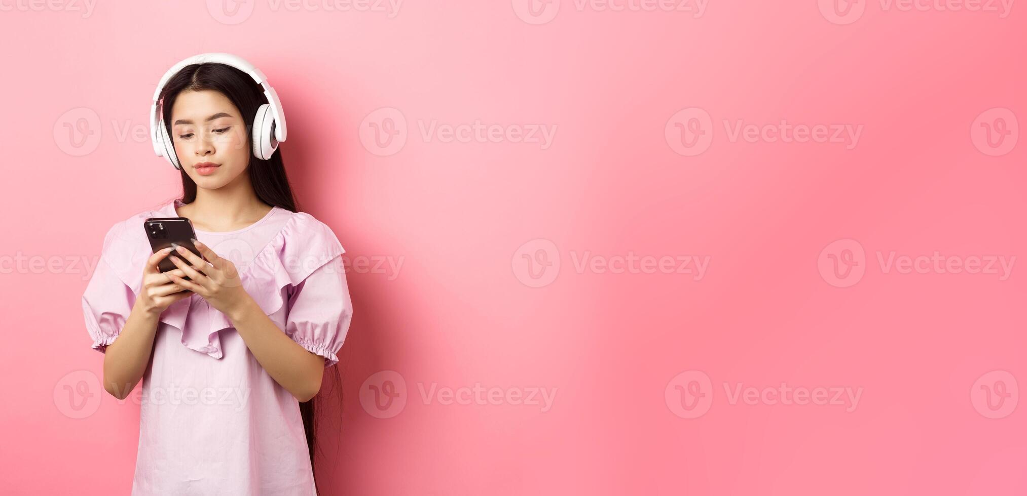 Young girl listening music in headphones and chatting on mobile phone, standing in dress against pink background photo