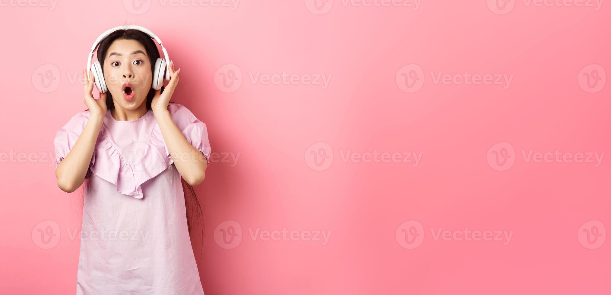 Excited asian girl hear awesome song, listen music in headphones and look amazed, standing on pink background photo