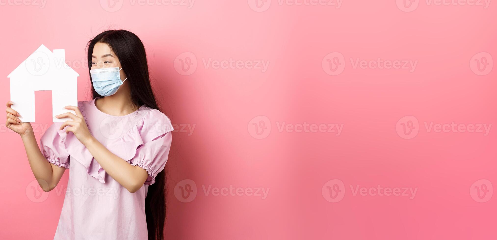 Real estate and pandemic concept. Excited asian girl in medical mask looking pleased at paper house cutout, buying property, standing on pink background photo