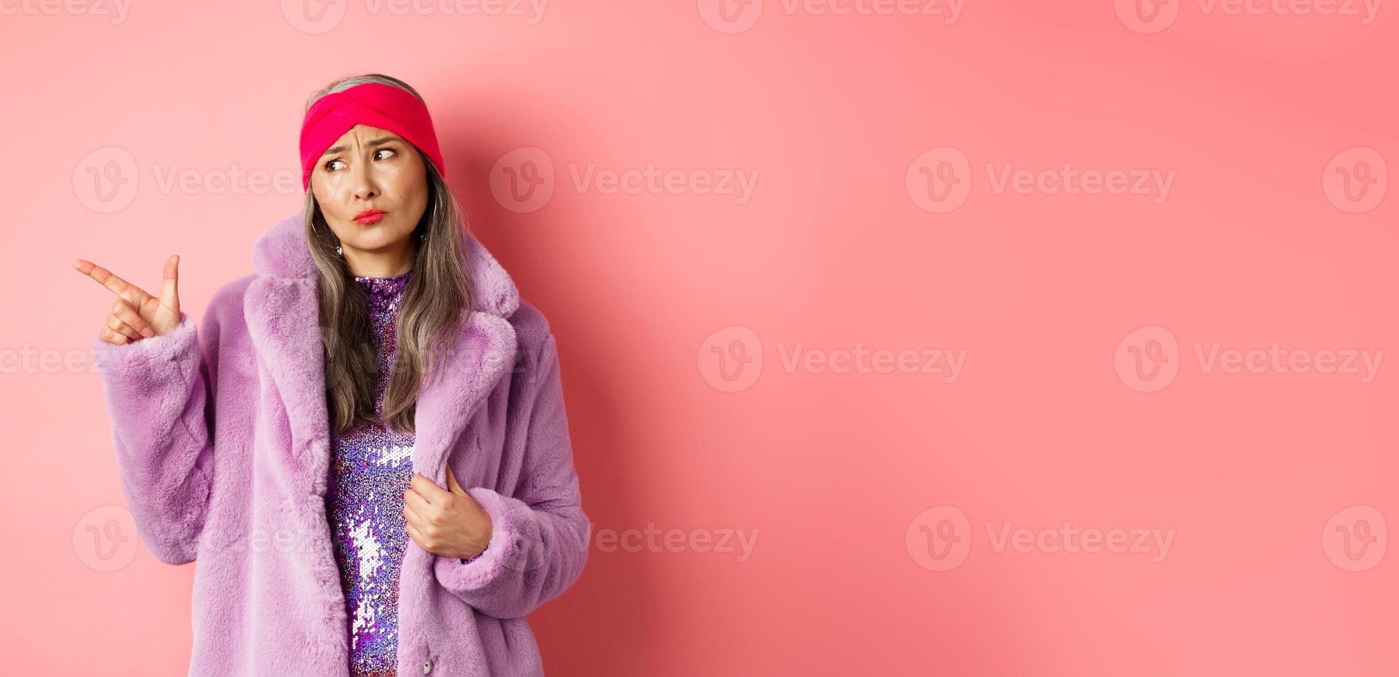 Fashion and shopping concept. Skeptical middle-aged asian woman in purple fur coat looking and pointing finger left with displeased, sad expression, standing over pink background photo