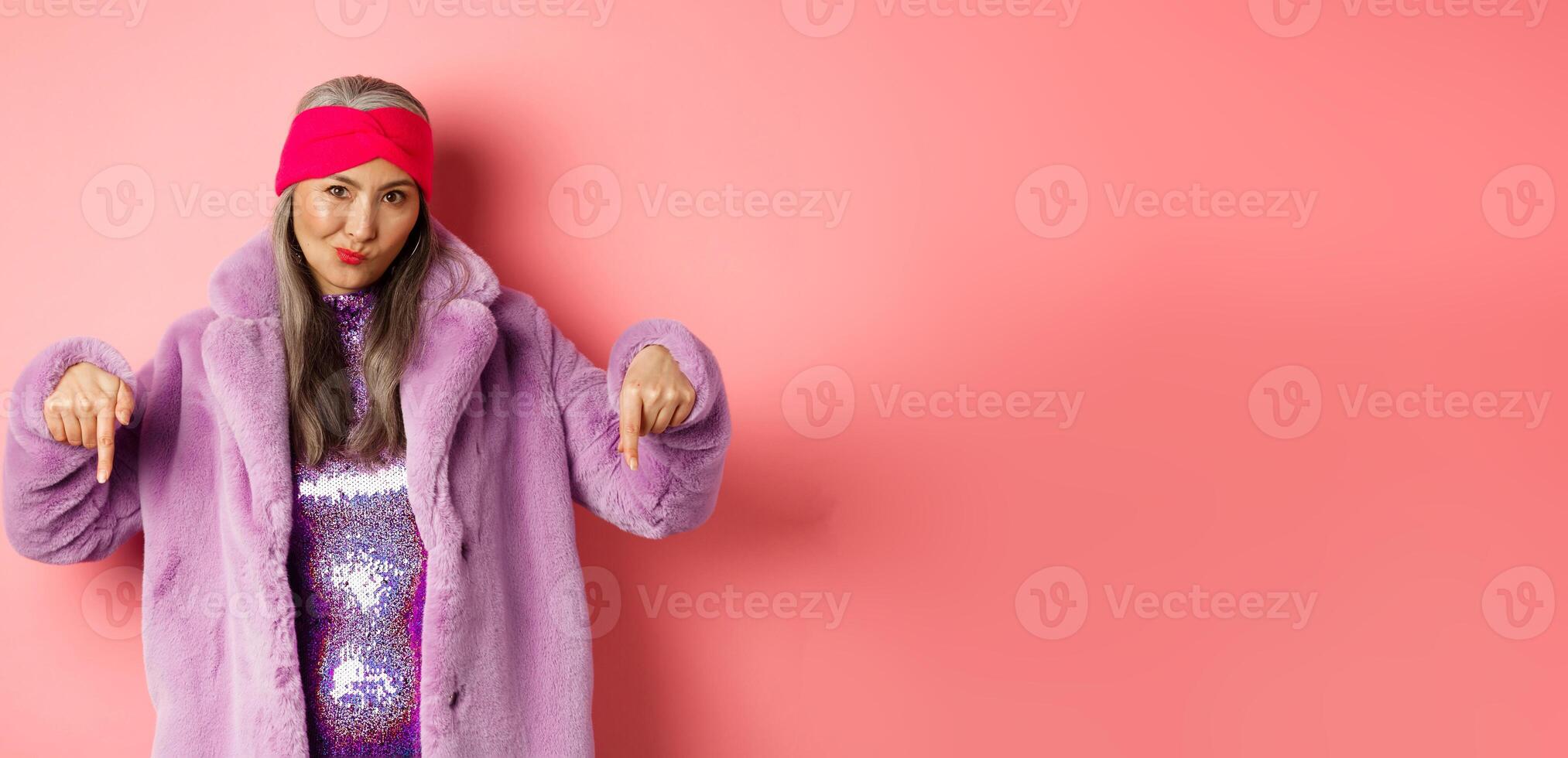 Fashion and shopping concept. Cool asian senior woman pointing fingers down, check out promo offer, looking sassy at camera, pink background photo