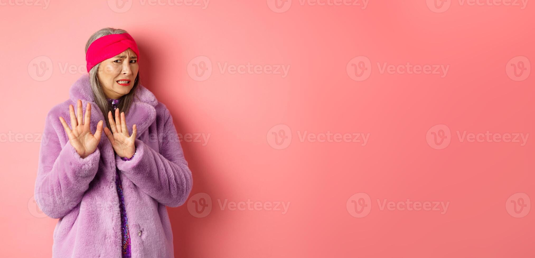Portrait of stylish asian senior woman in purple winter coat, cringe from something disgusting, frowning and turn away awkward, showing stop gesture, refusing on pink background photo