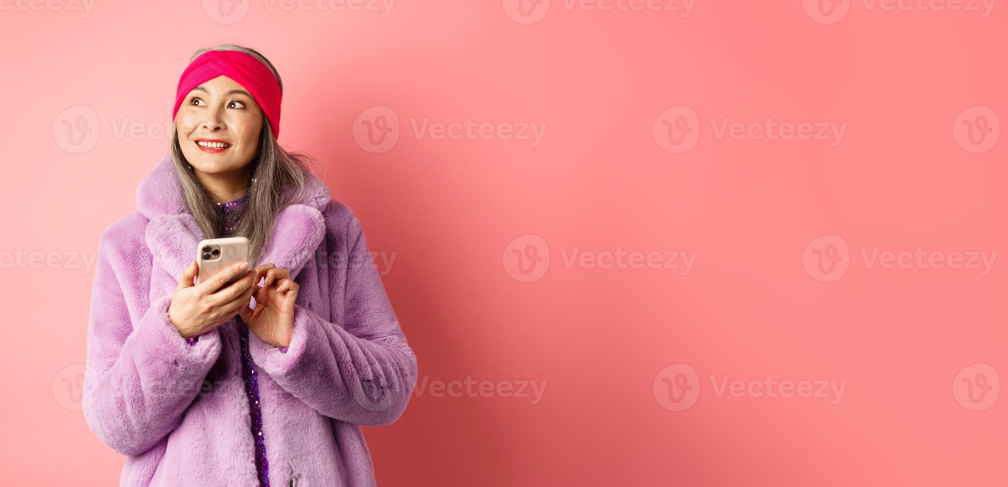 Online shopping and fashion concept. Stylish asian senior woman using smartphone, looking aside with thoughtful face, standing against pink background photo