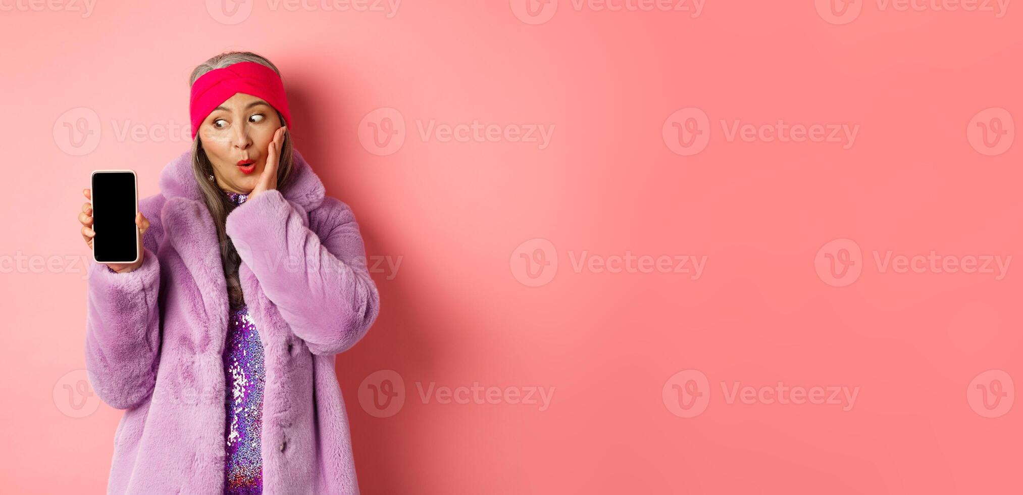 Valentines day shopping concept. Beautiful asian senior woman showing smartphone screen and looking at display excited, checking out online offer, pink background photo