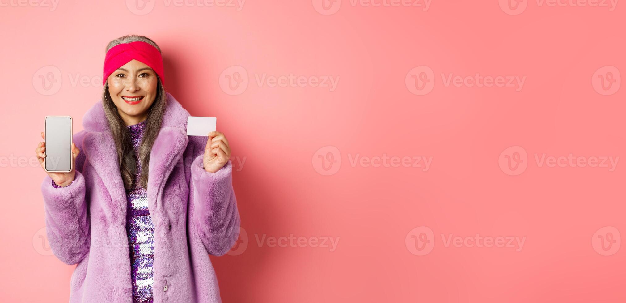 Online shopping and fashion concept. Fashionable asian senior woman showing black screen of smartphone and plastic credit card, smiling happy, pink background photo