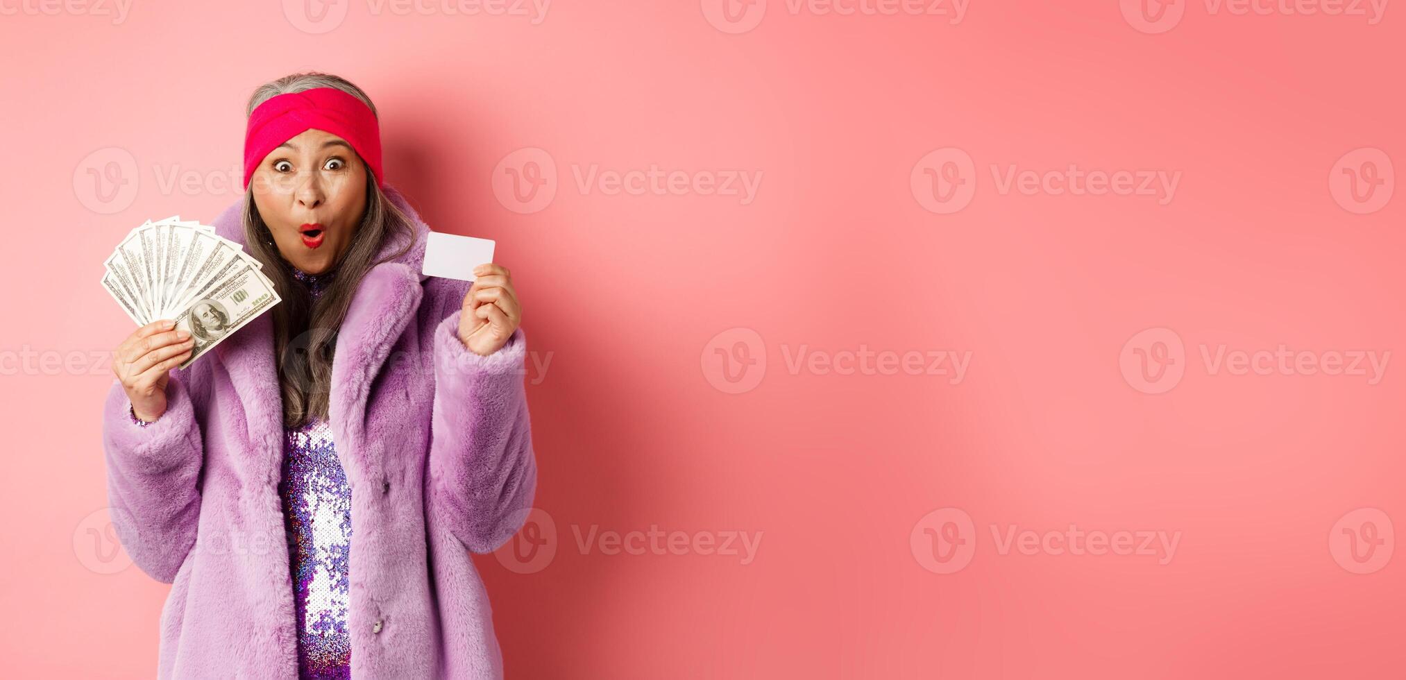 Shopping and fashion concept. Happy senior asian woman holding money in dollars and plastic card, looking amazed and ecstatic, pink background photo