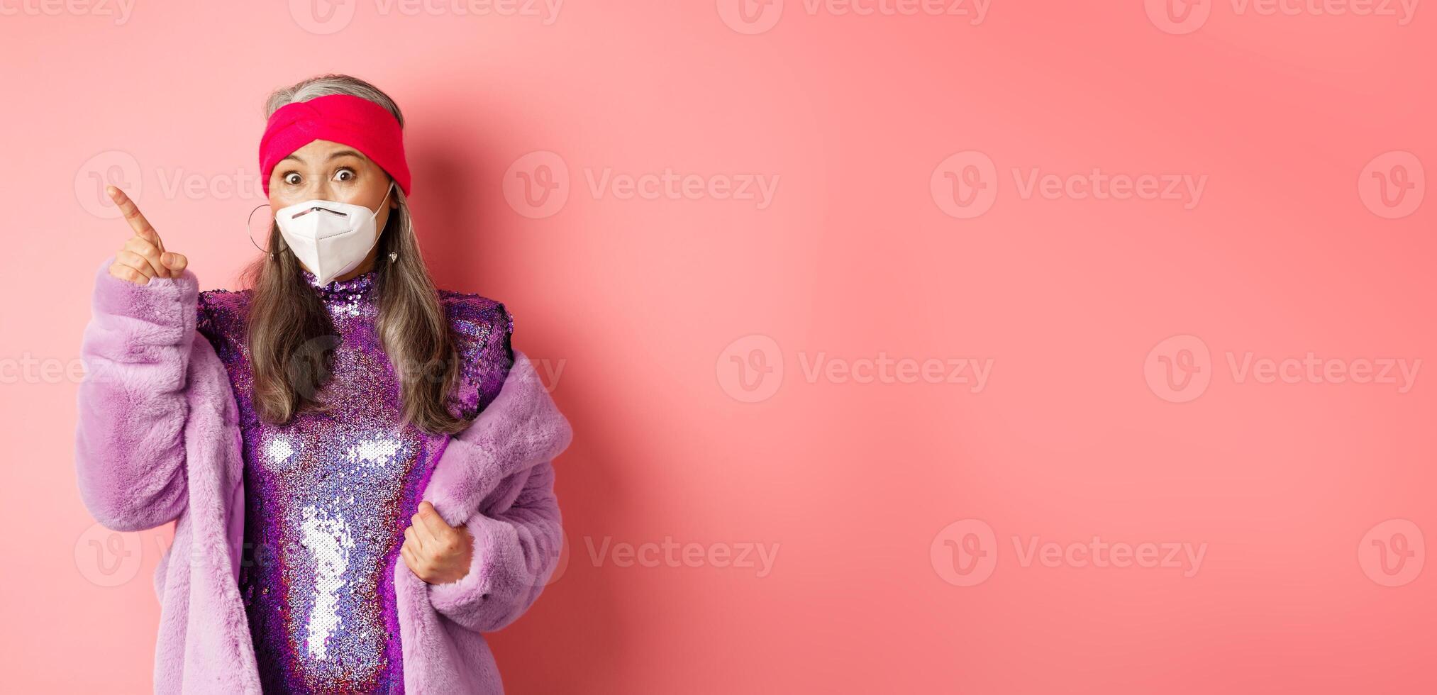 Covid and fashion concept. Amazed asian senior woman in glittering dress and face mask, pointing finger upper left corner, showing promo offer, pink background photo