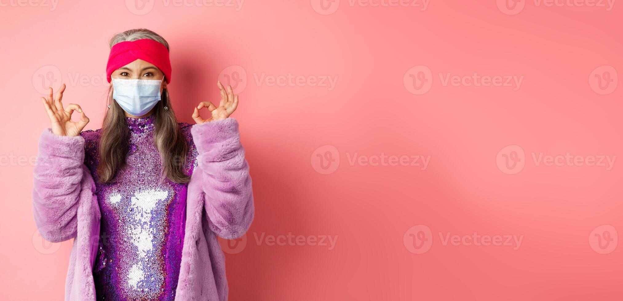 Covid-19, social distancing and fashion concept. Fashionable asian senior woman in face mask and glitter dress, showing okay signs in approval, pink background photo
