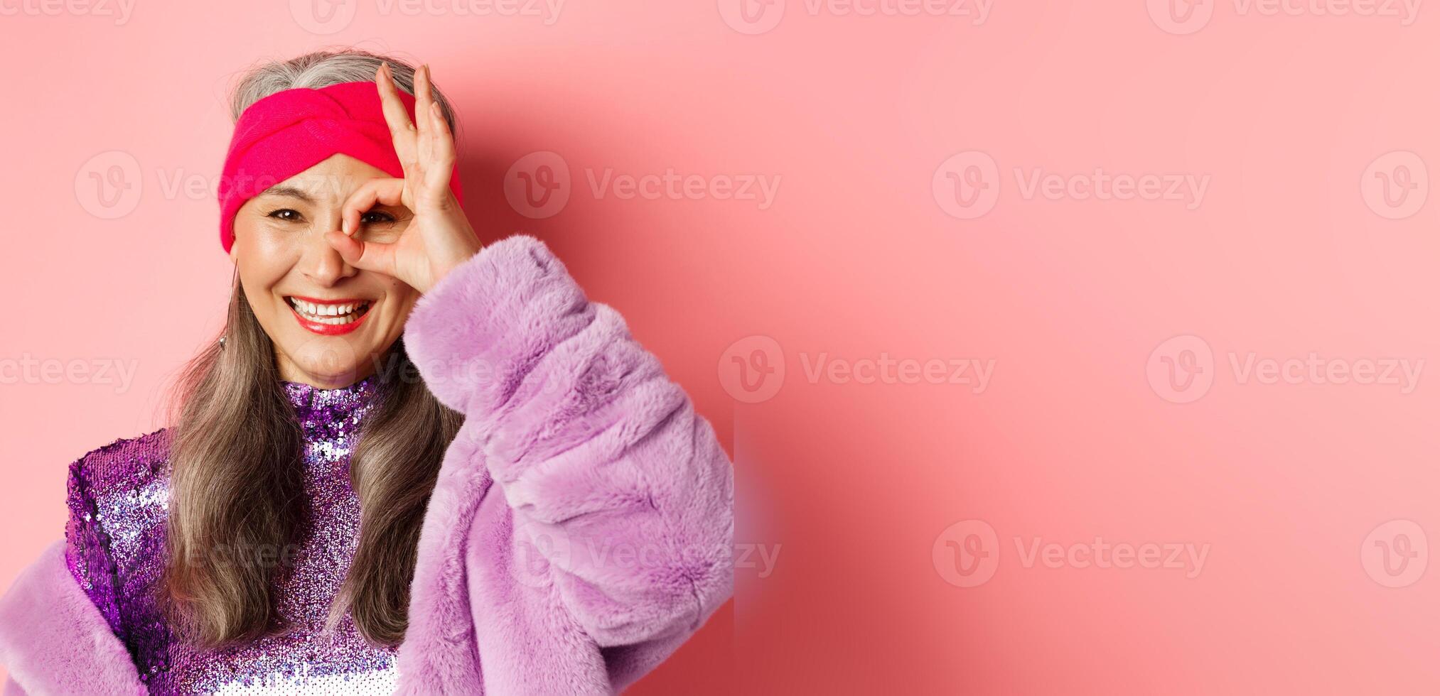 Fashion. Close-up of fashionable asian senior woman smiling, showing OK sign over eye and looking happy at camera, standing over pink background photo