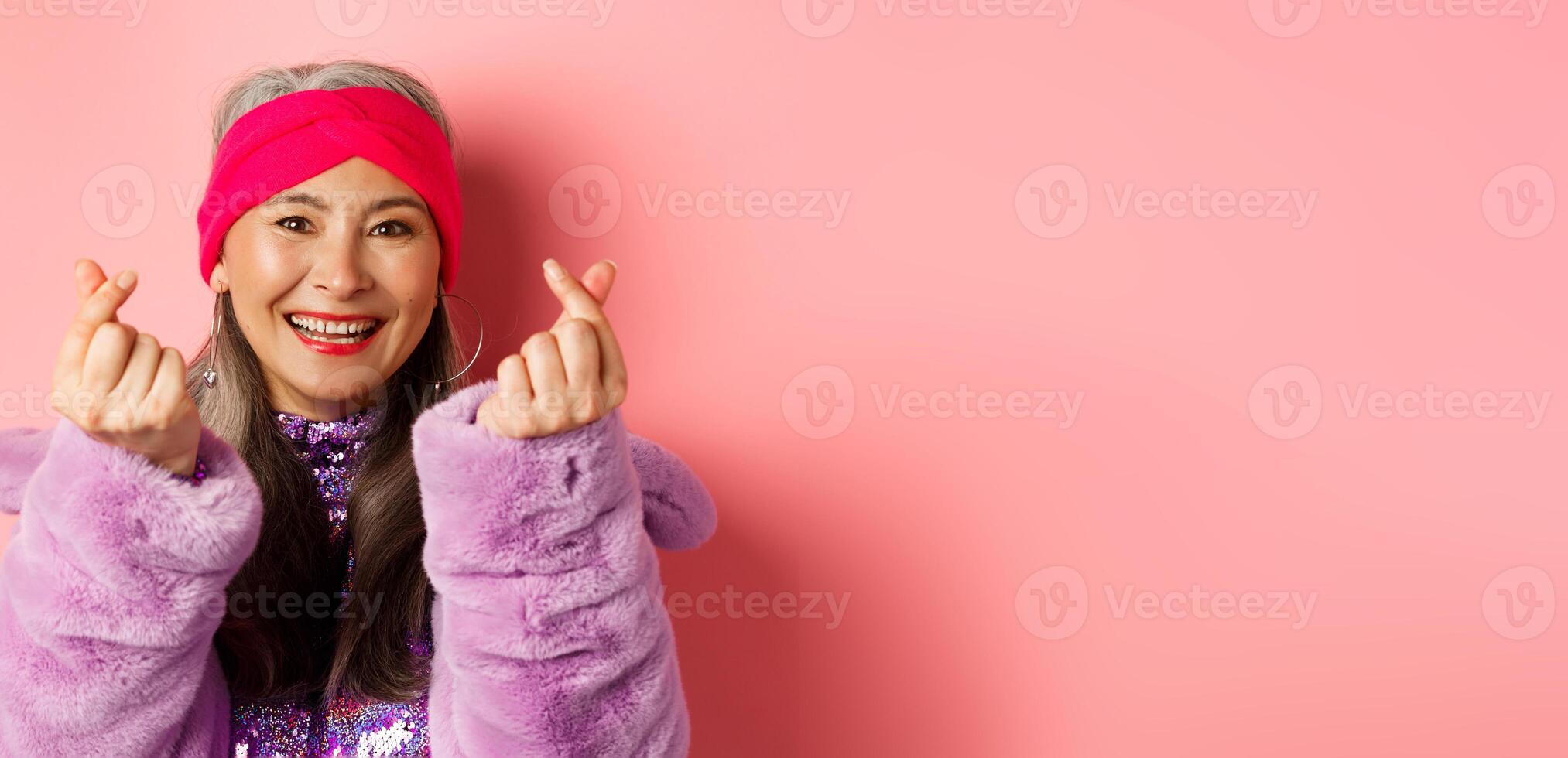 Fashion. Beautiful and fashionable asian senior woman smiling, showing korean heart gesture and looking happy, standing over pink background photo