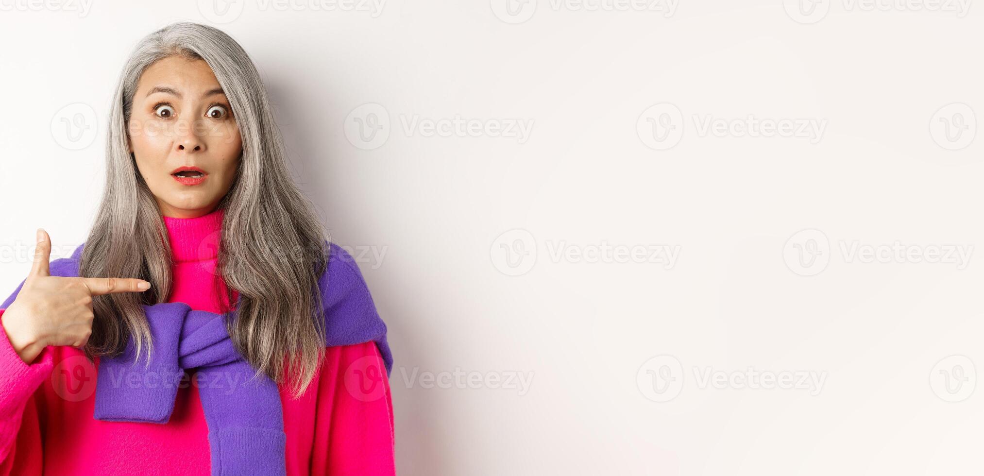 Confused asian senior woman with grey hair gasping startled, pointing at herself and looking at camera, standing over white background photo