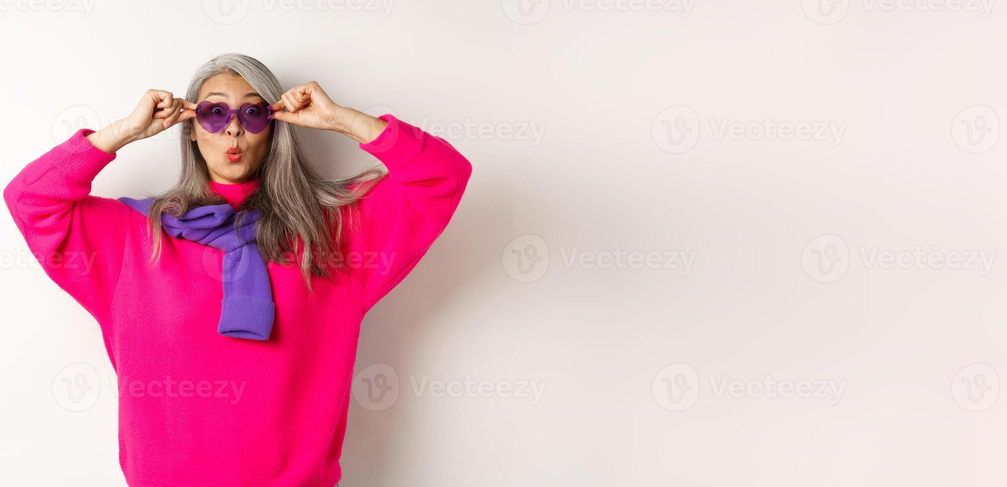 Fashionable asian senior woman in sunglasses and stylish pink sweater pucker lips, raising eyebrows and looking surprised at camera, white background photo
