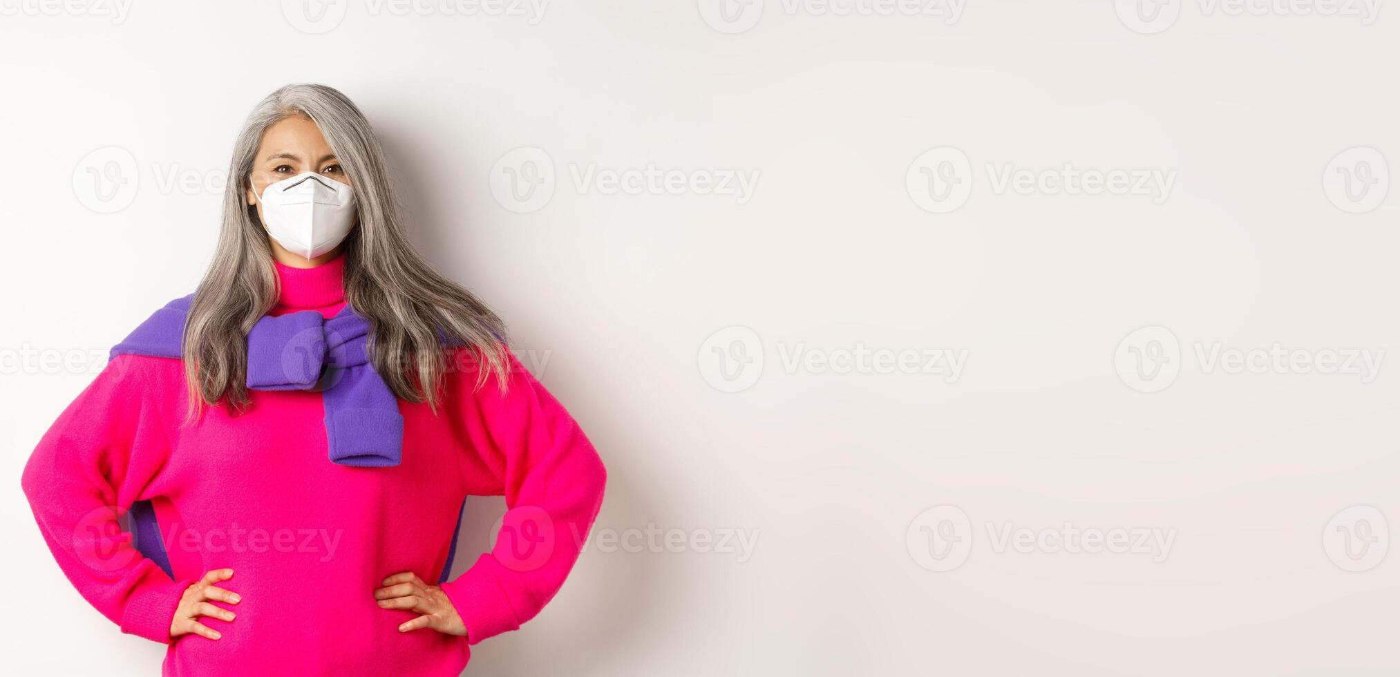 Covid, pandemic and social distancing concept. Confident and cheerful asian senior woman in respirator looking upbeat, smiling and holding hands on waist, white background photo