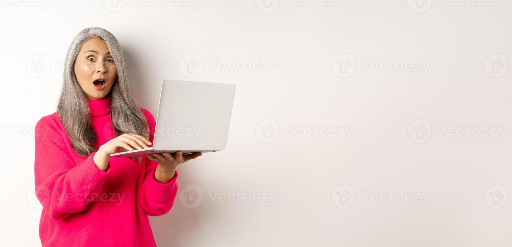 Surprised and impressed asian grandmother staring shocked at camera after reading news on laptop screen, standing over white background photo