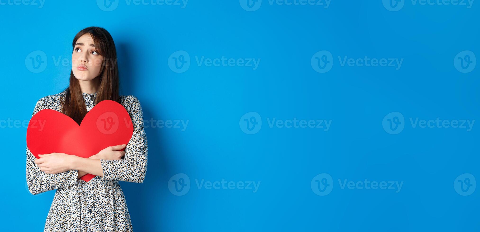 Valentines day. Sad heartbroken girl sobbing, looking aside at empty space and crying upset, hugging big red heart, feeling lonely without lover, standing alone on blue background photo