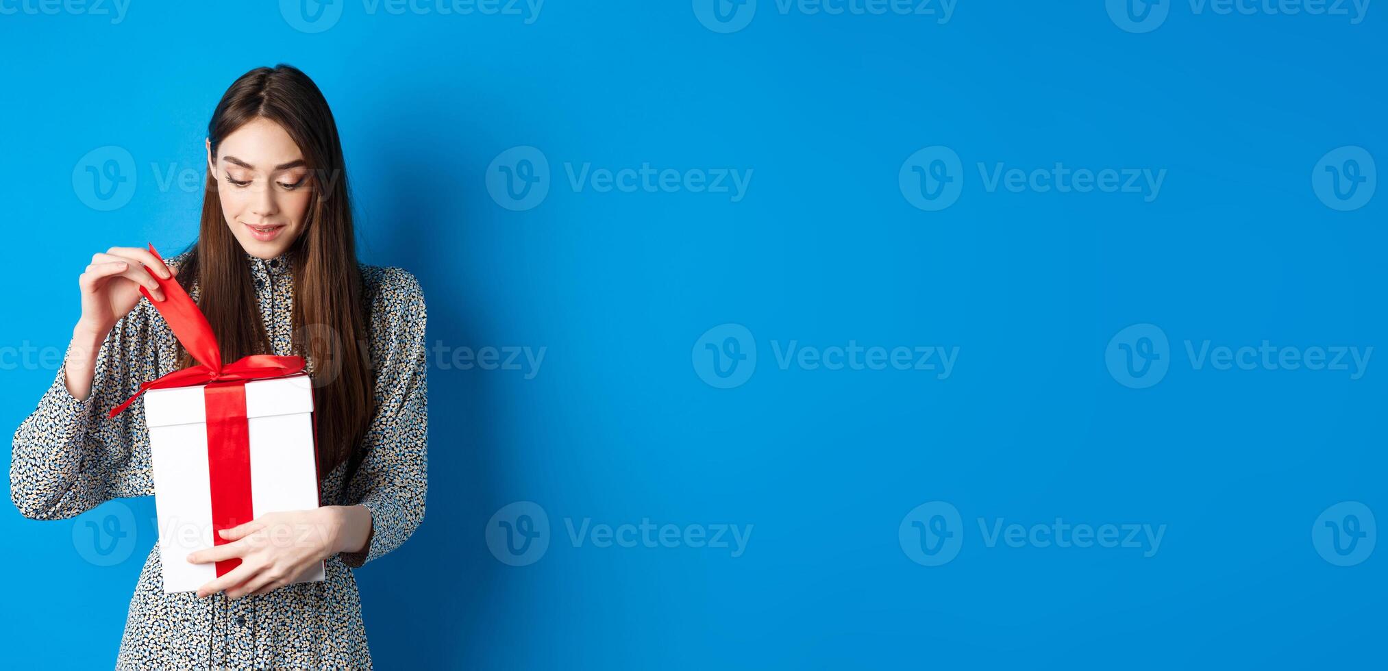 Valentines day. Cute young woman open box with gift, take-off ribbon from present and smiling intrigued, standing on blue background photo