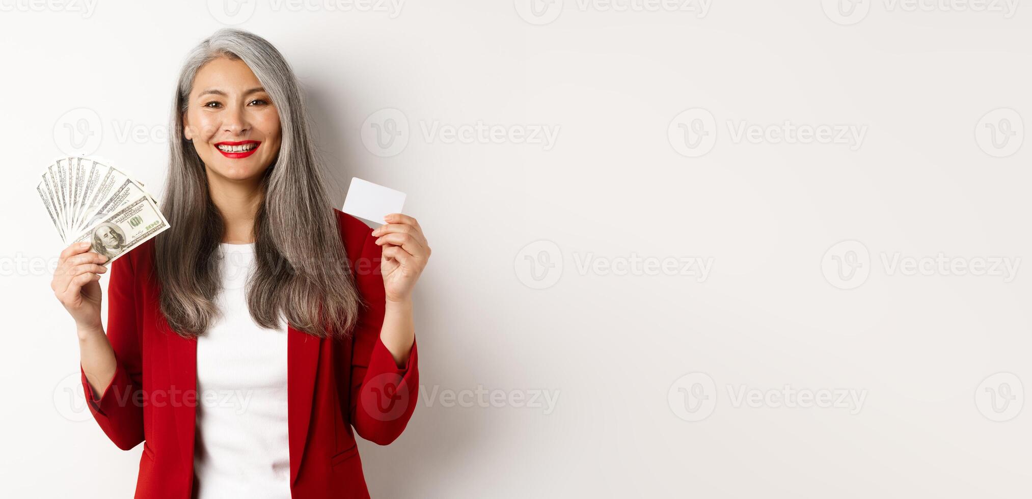 Successful asian senior businesswoman showing money in dollars and plastic card, smiling happy at camera, wearing red blazer and make-up photo