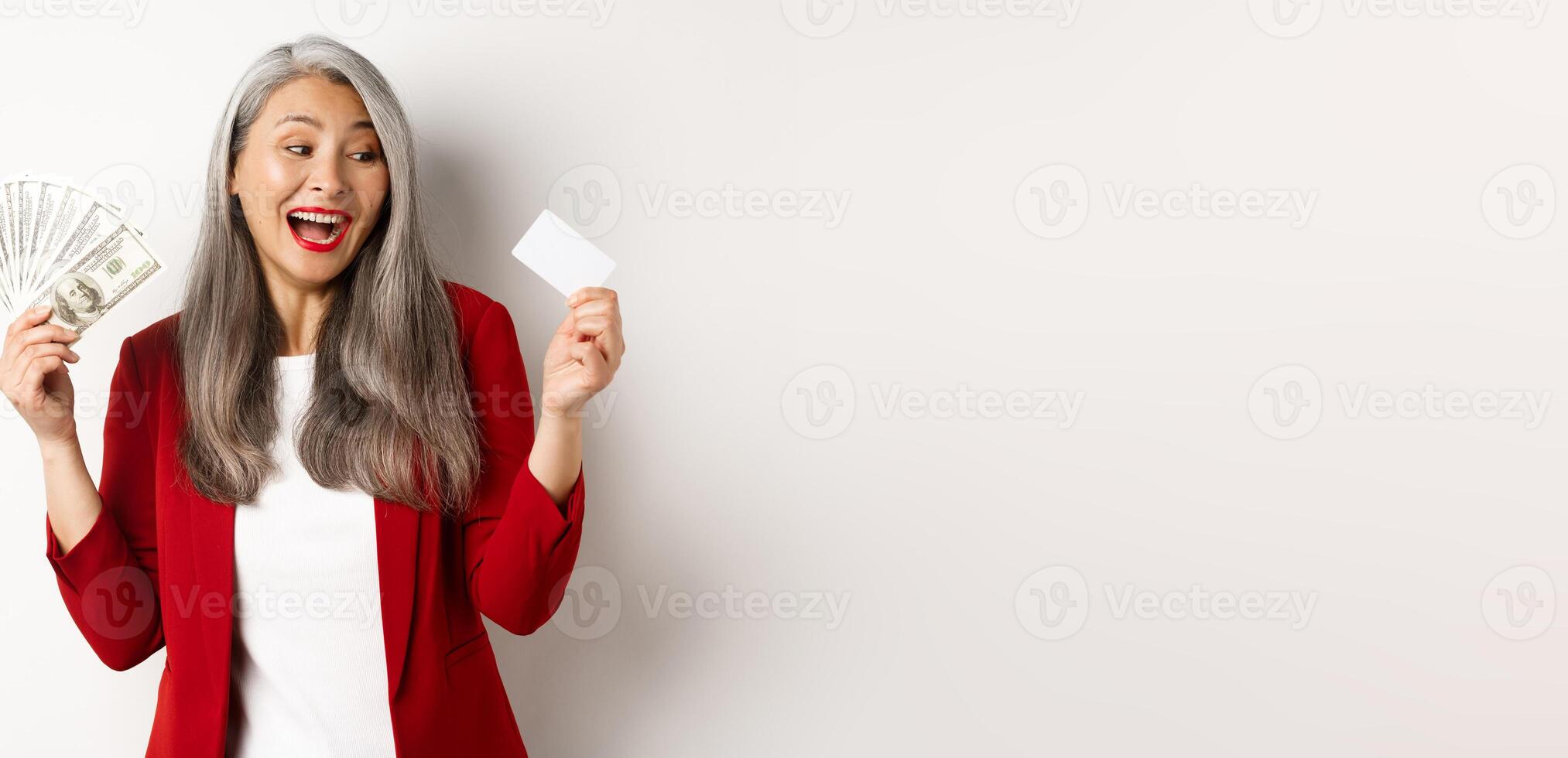 Cheerful asian senior woman earn money, showing dollars in cash and looking at plastic credit card, smiling excited, standing over white background photo