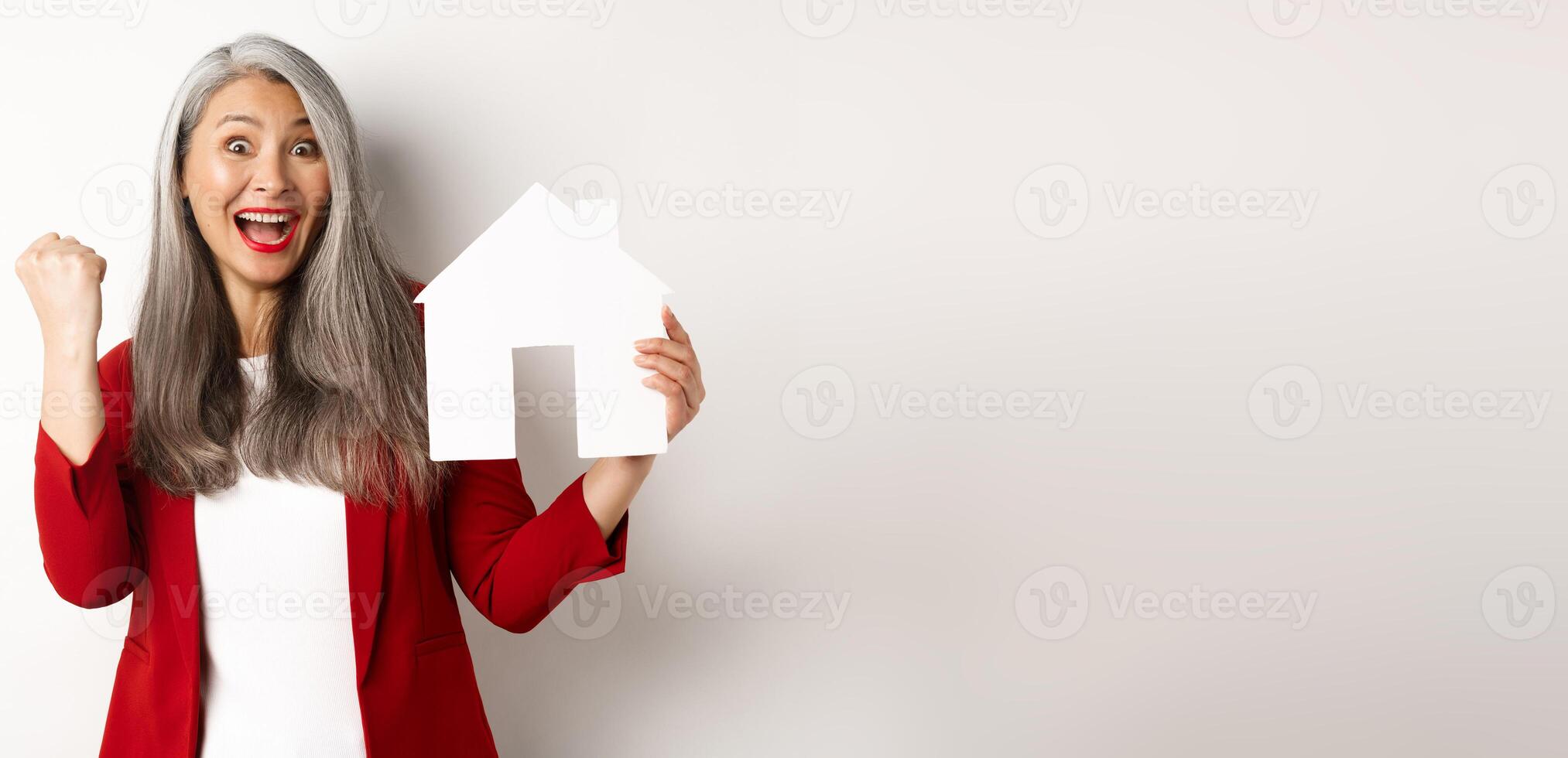 Happy asian grandmother showing paper house cutout and fist pump gesture, scream yes with joy, buying property, standing over white background photo