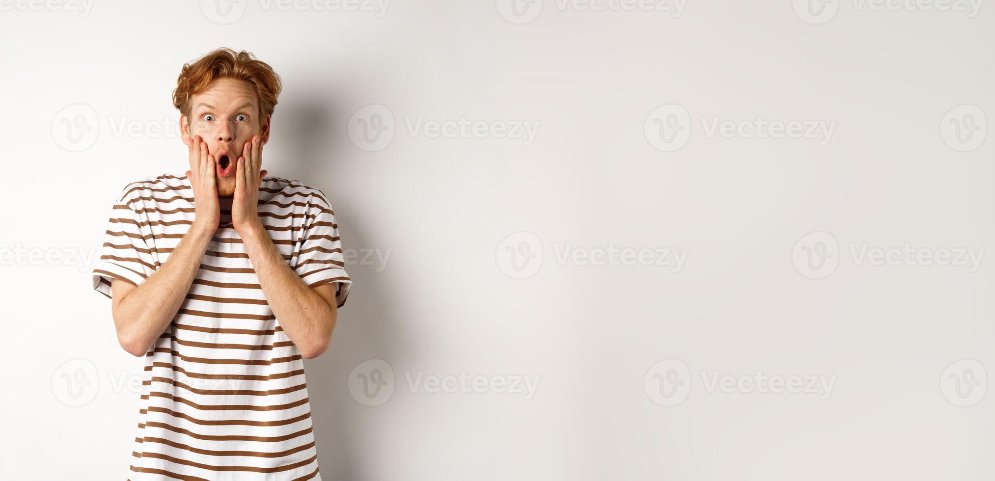 Impressed young man with ginger hair, gasping and staring shocked at camera, express complete disbelief and amazement, white background photo