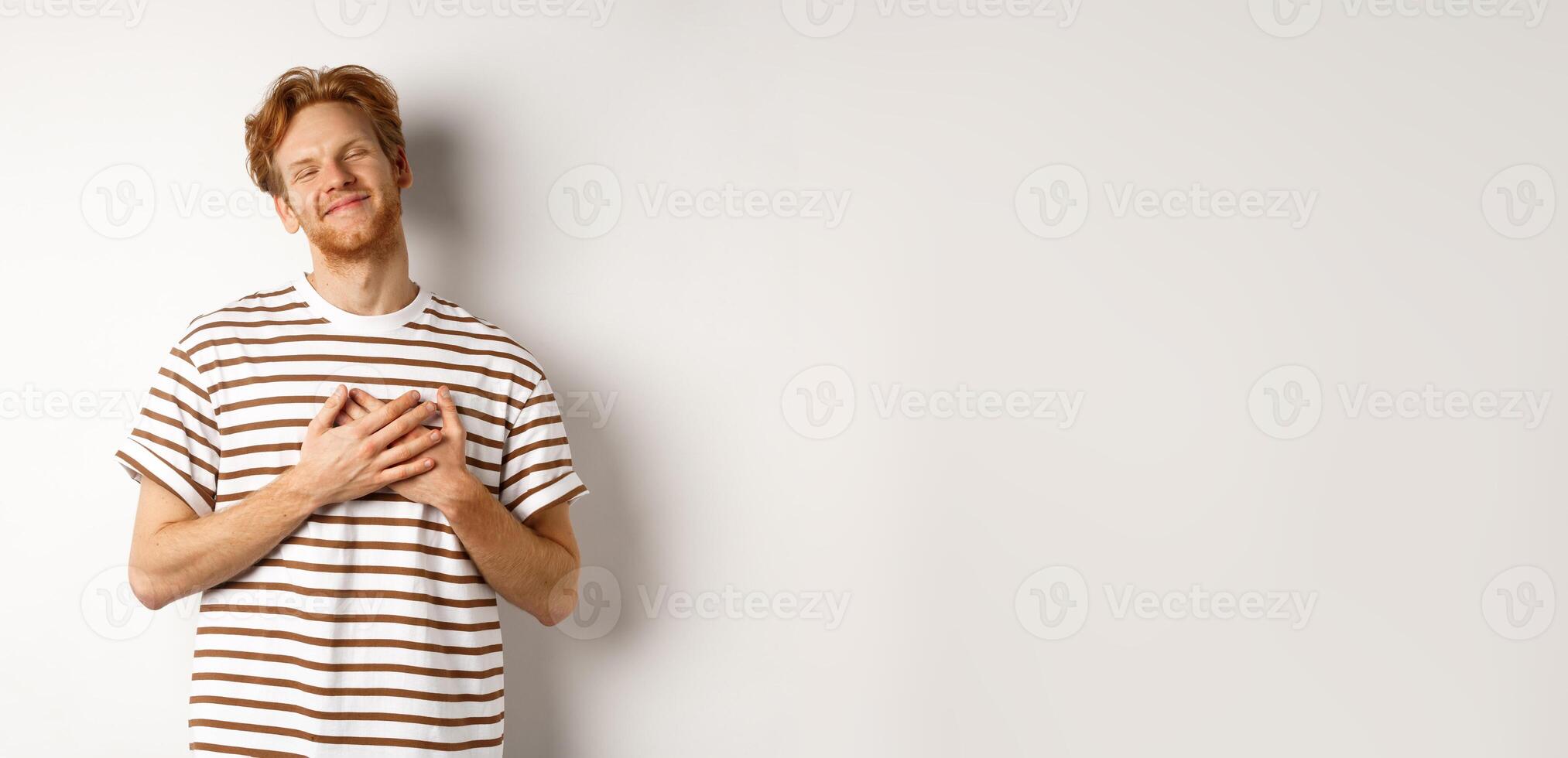 Happy young man with ginger hair appreciate gift, holding hands on heart and smiling, thanking you, standing grateful against white background photo