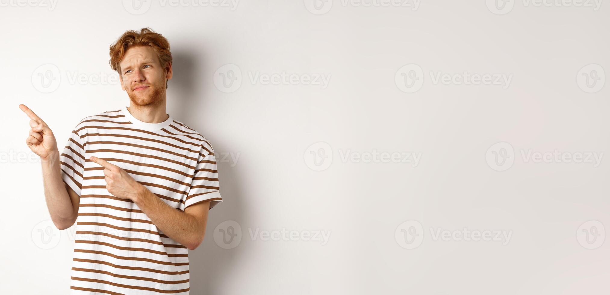 Doubtful young man with bristle and red hair looking, pointing fingers left at something average, staring skeptical at promo, white background photo