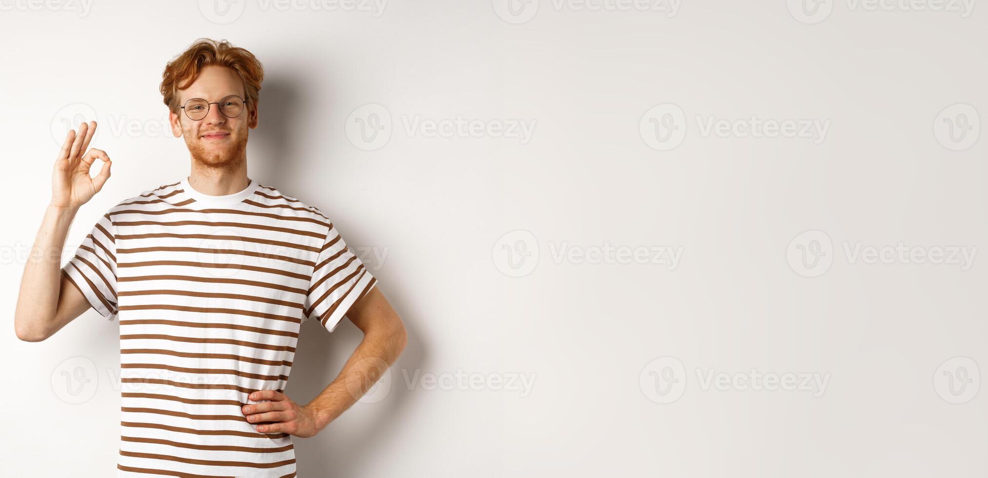 Proud and happy man with red hair and glasses smiling, showing OK sign, praising something excellent, saying yes or good, standing over white background photo