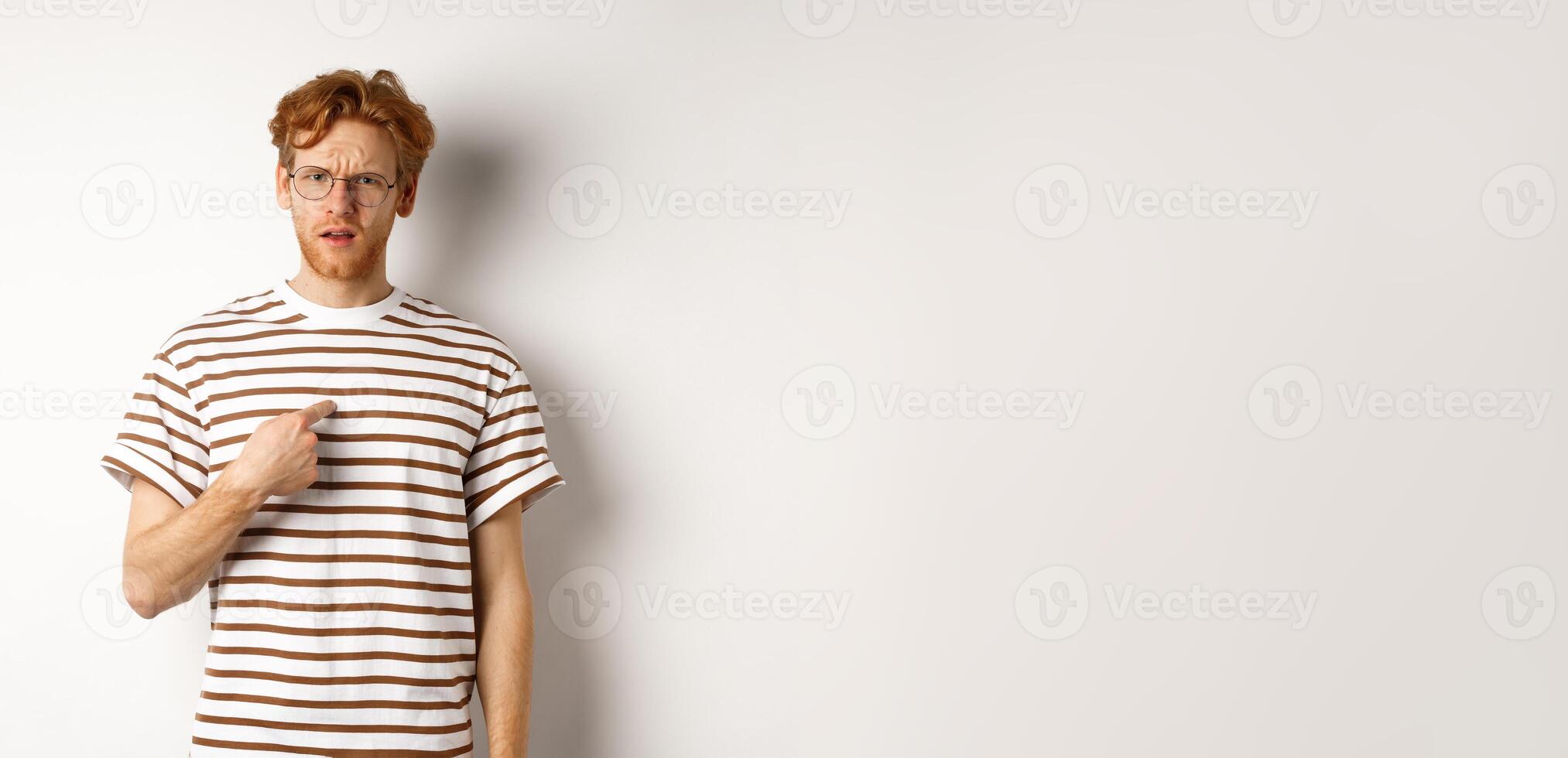 Confused redhead man in glasses pointing at himself, looking questioned at camera, white background photo