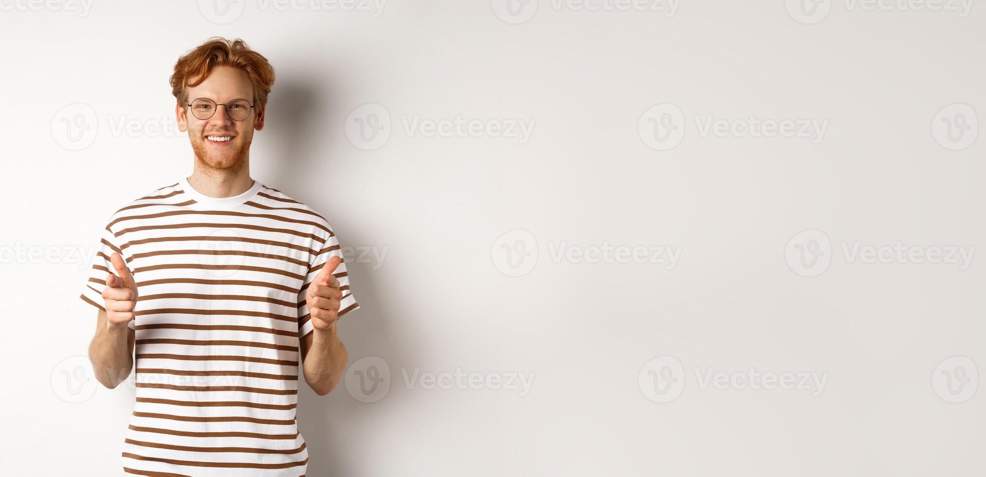 Young man with red hair and glasses pointing fingers at camera and smiling, need you, standing over white background photo