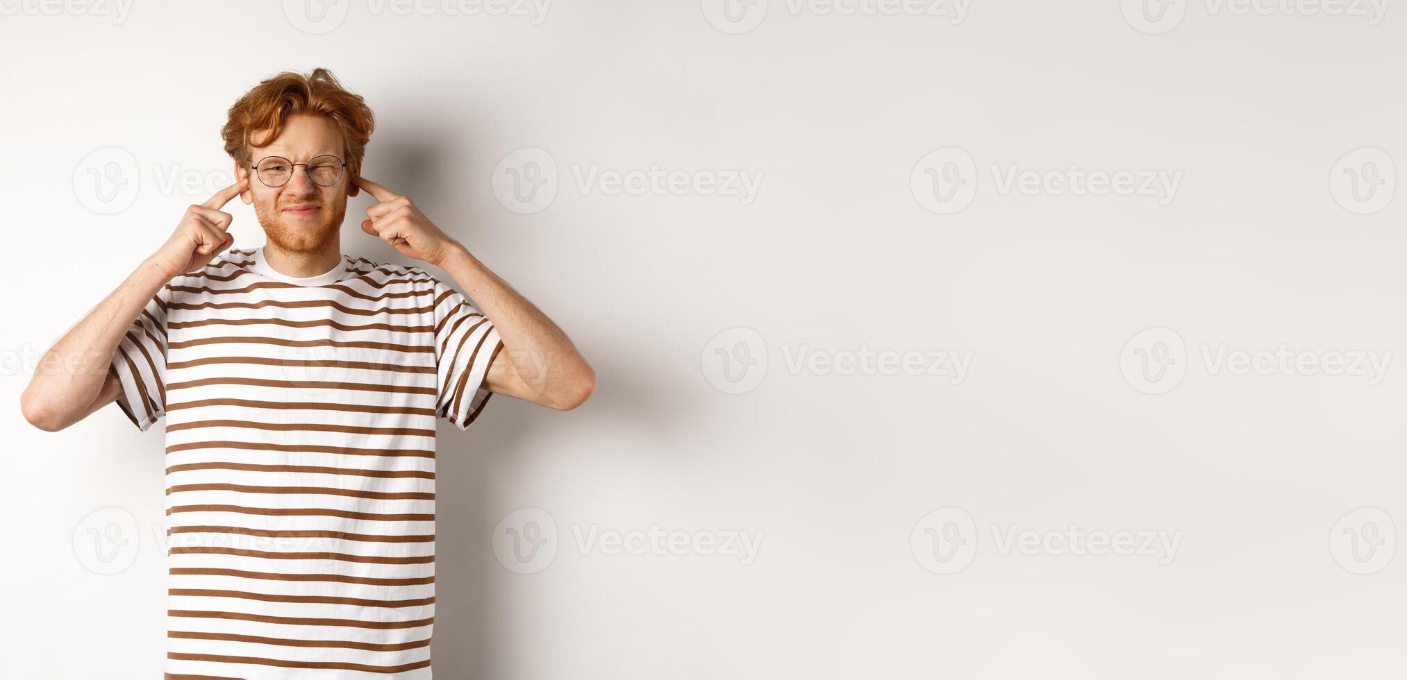 Annoyed young man complaining on loud noise, shut ears and grimacing from discomfort, standing over white background photo