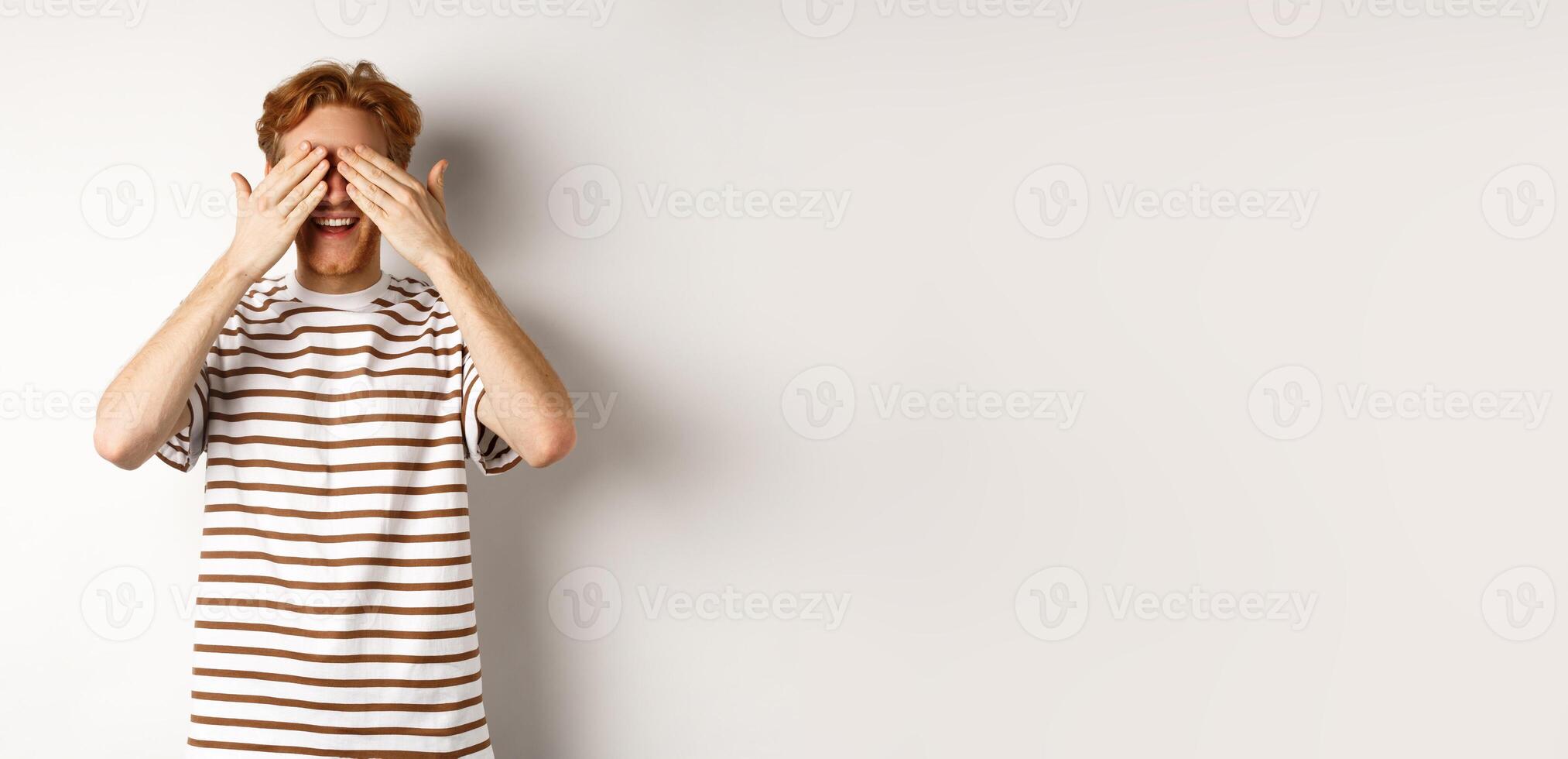 Smiling man cover eyes and waiting for surprise, standing in t-shirt against white background photo