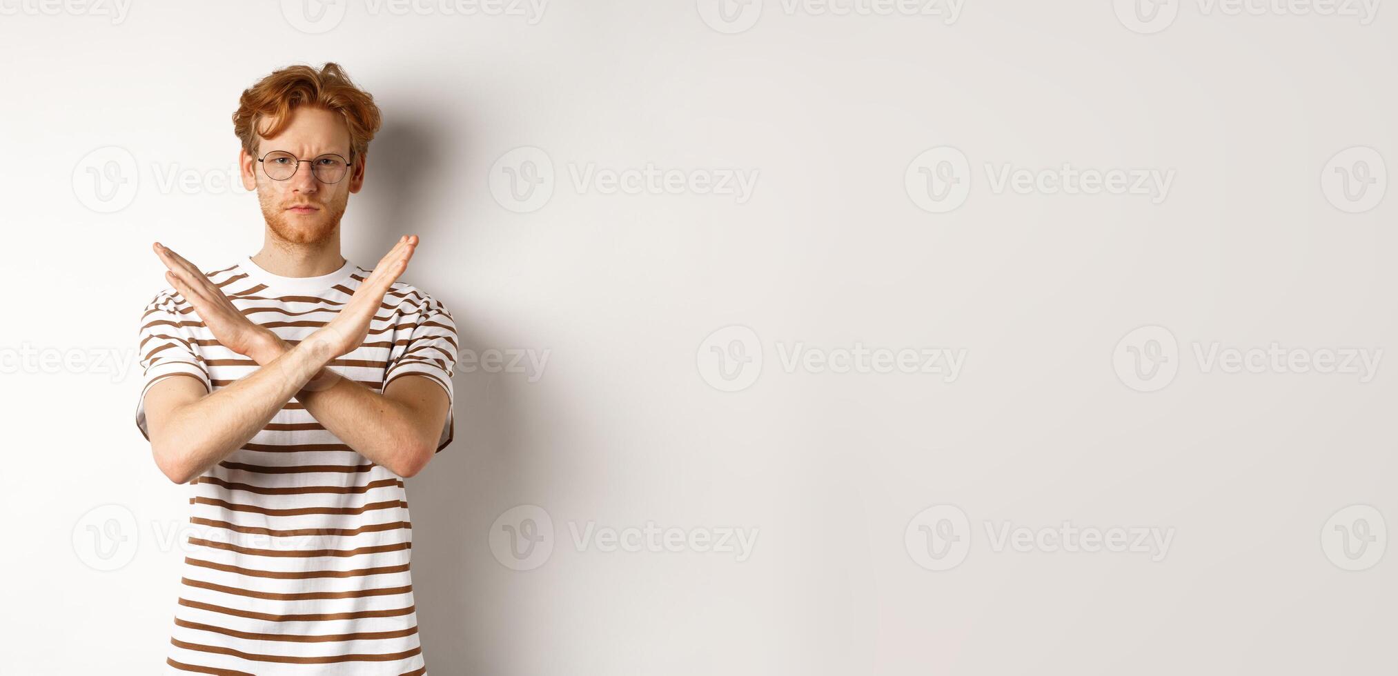 Serious young man with ginger hair frowning, showing stop gesture, making cross to prohibit something bad, disagree with you, standing over white background photo