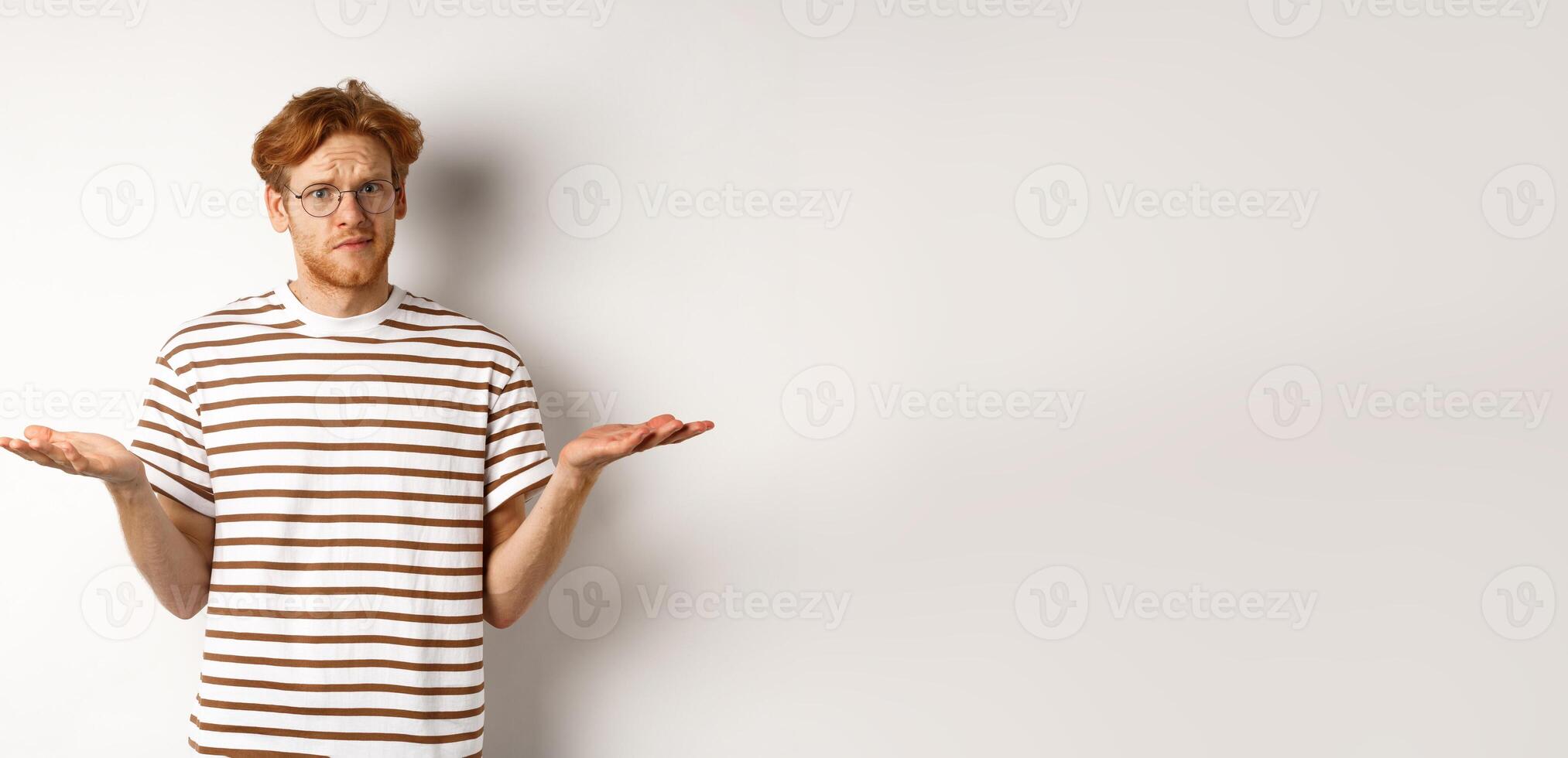 Confused boyfriend with red hair shrugging, spread hands sideways and staring at camera puzzled, asking what, stnading over white background photo
