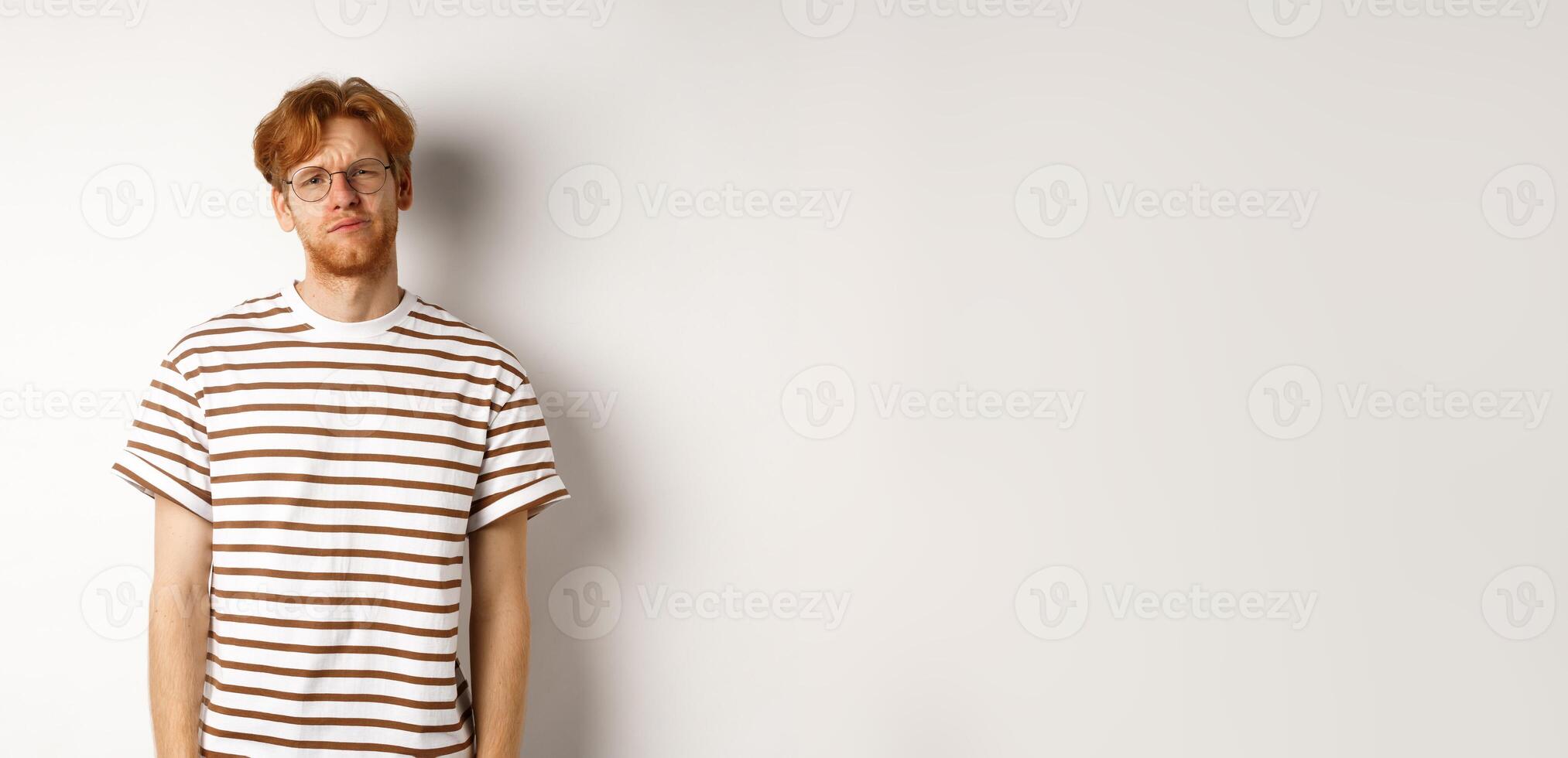 Reluctant and unamused redhead young man staring at camera, looking tired or displeased, standing over white background photo