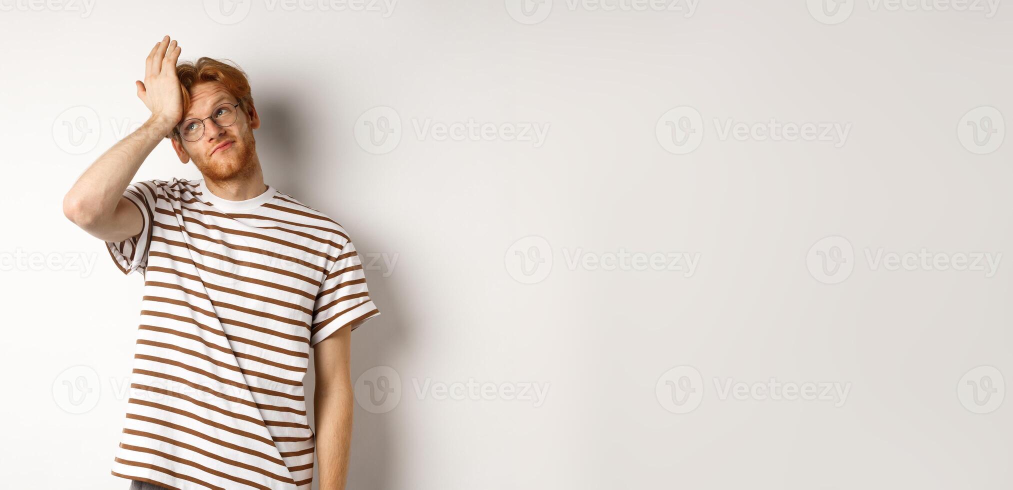 Tired young man with red hair and glasses, roll eyes and making facepalm bothered, standing over white background photo
