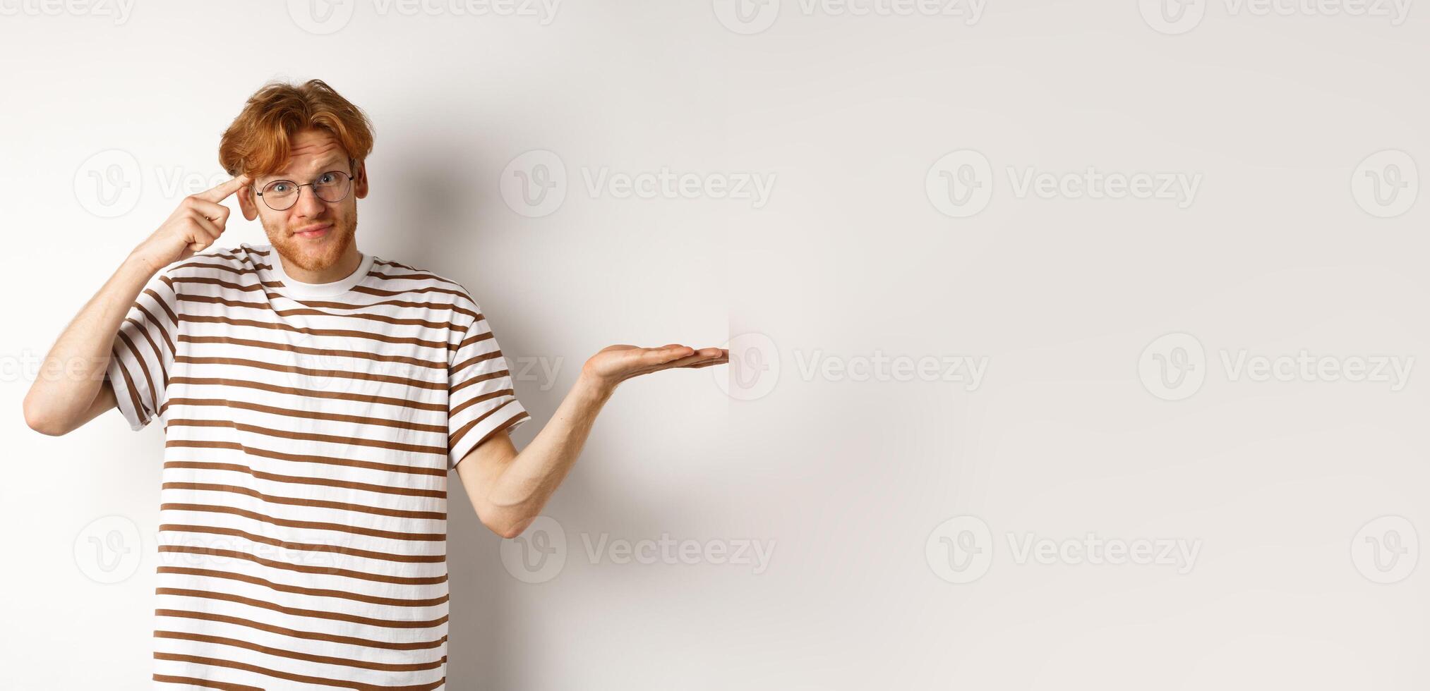Are you stupid. Confused redhead guy in glasses shrugging, pointing at head and staring skeptical at camera, white background photo